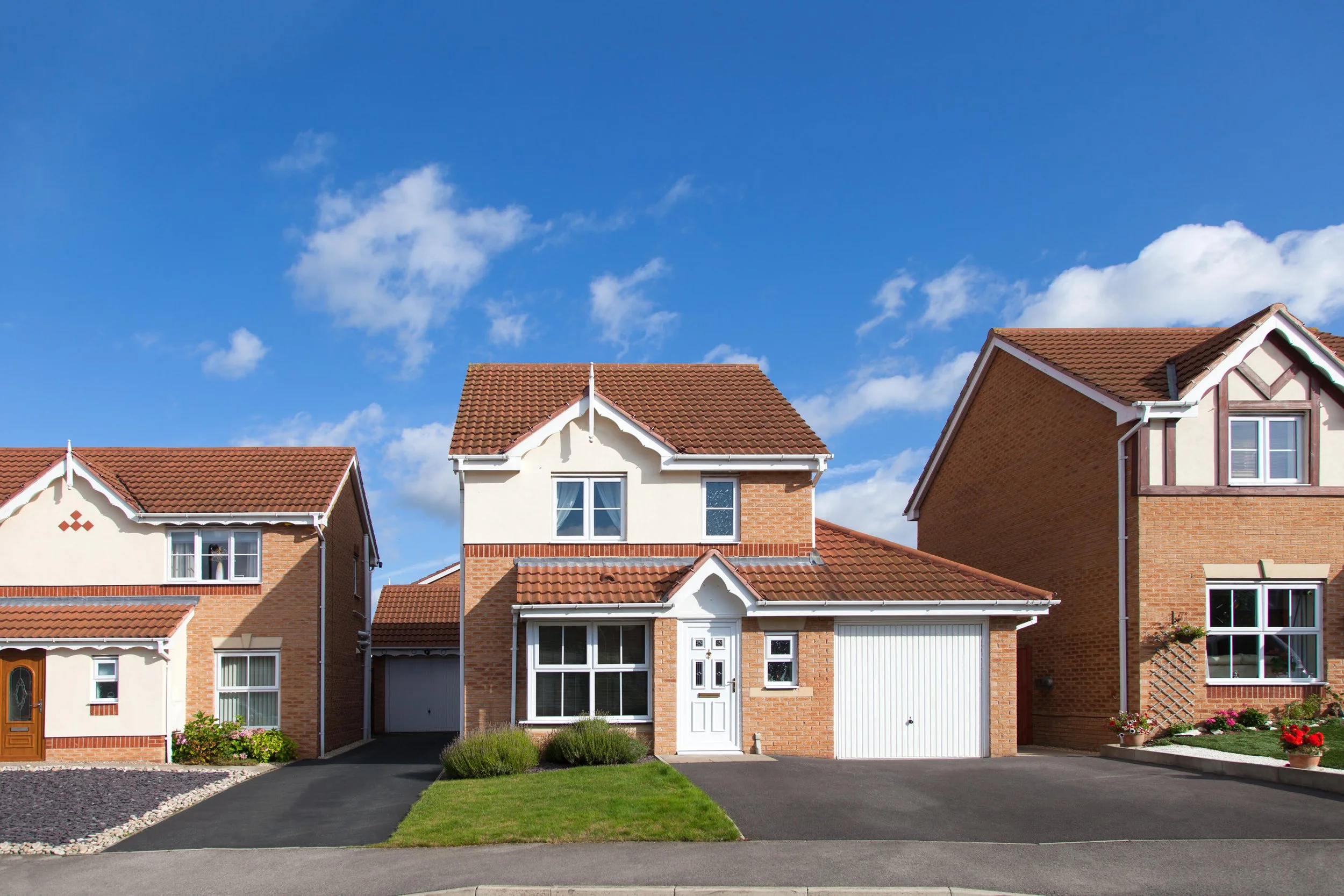 Front view of a modern two-story brick house with a white front door, attached garage, and landscaping, under a blue sky with scattered clouds.