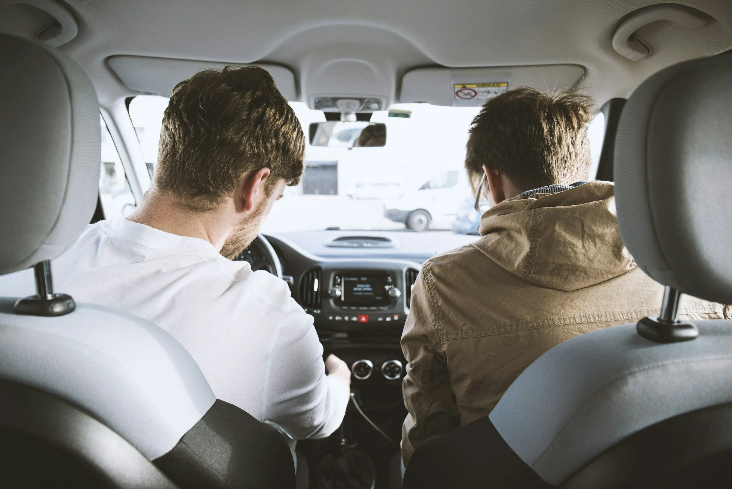 Two men are sitting in the front seats of a car, looking ahead and talking to each other.