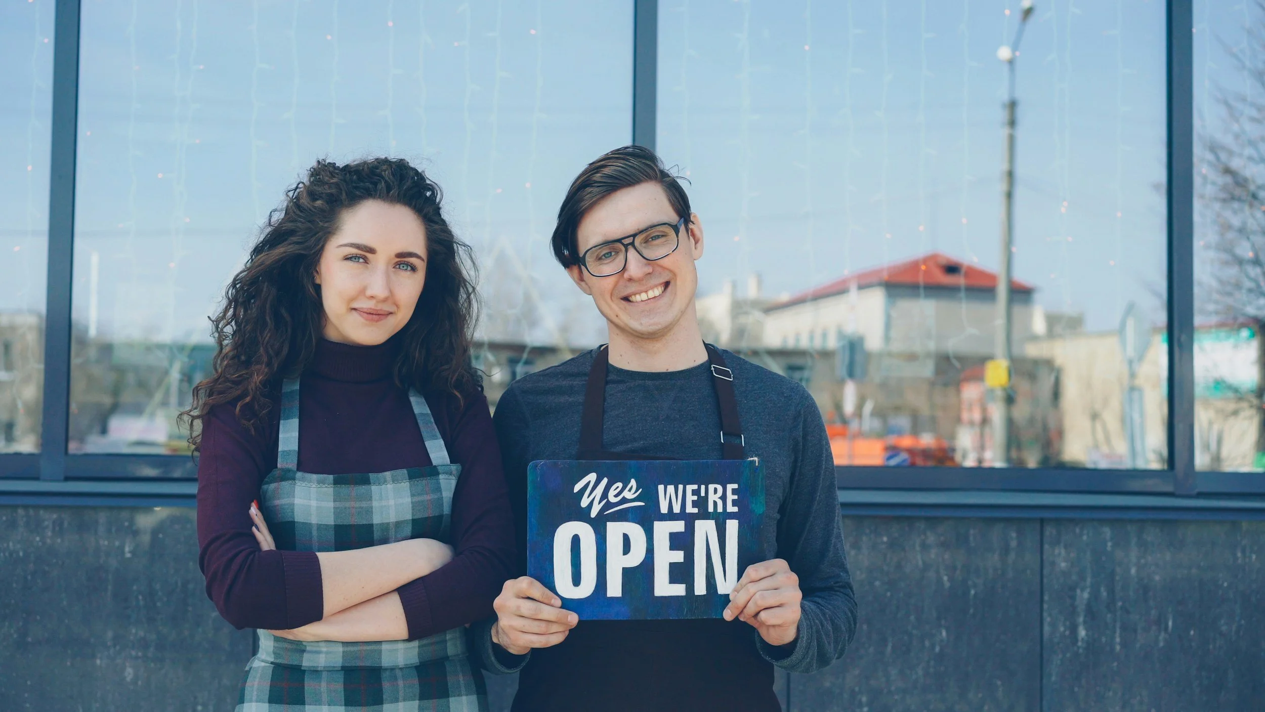 Two smiling young adults, a woman and a man, standing outside a store with a large glass window. The man is holding a sign that says "Yes We're Open." The woman is wearing a plaid apron and has crossed arms, while the man is wearing glasses and a dark shirt.