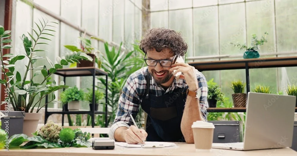 Young man talking on the phone while working at a desk filled with plants and office supplies in a greenhouse or plant nursery.
