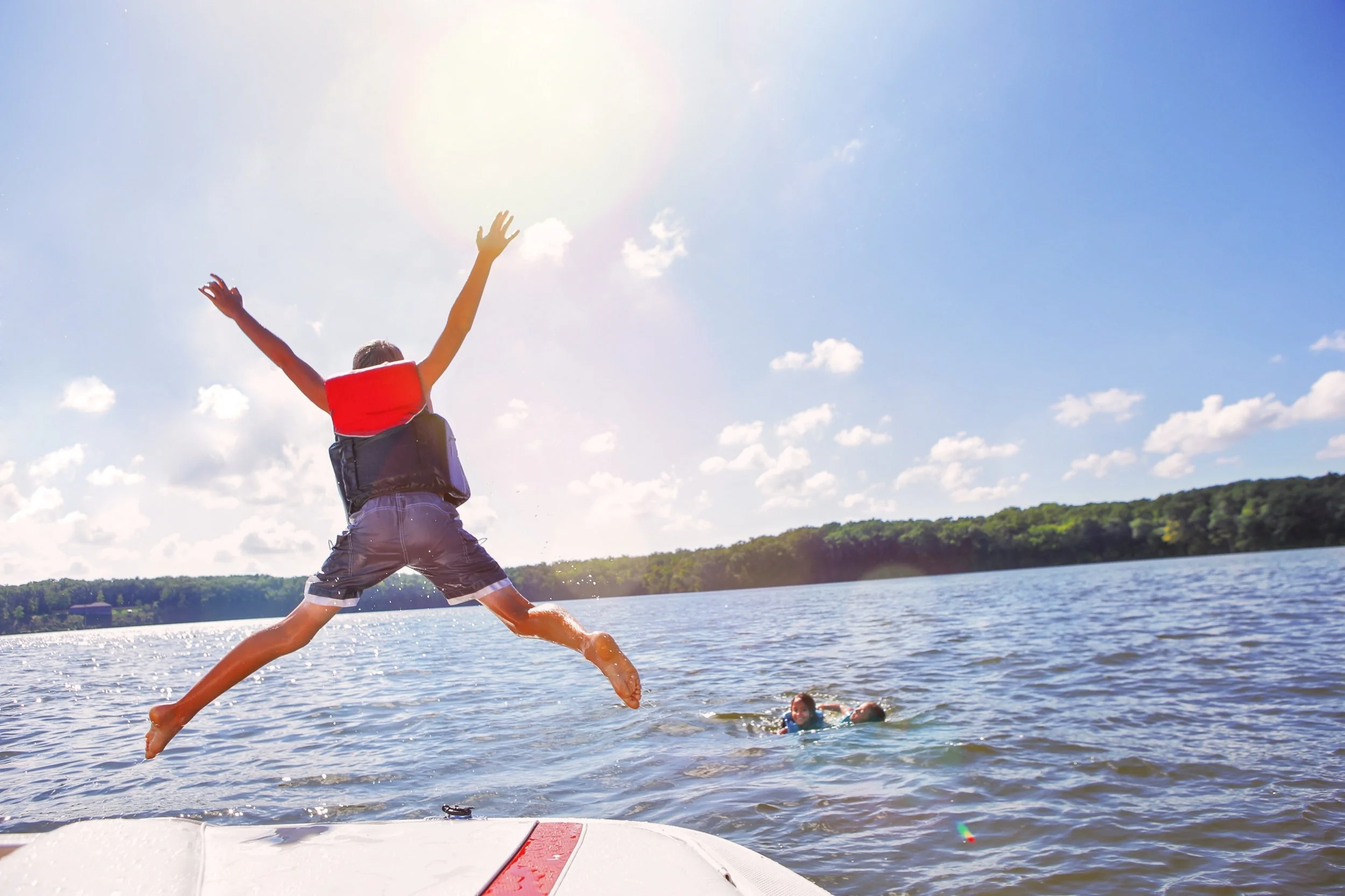 young boy jumping off boat with life jacket in the water with other kids playing