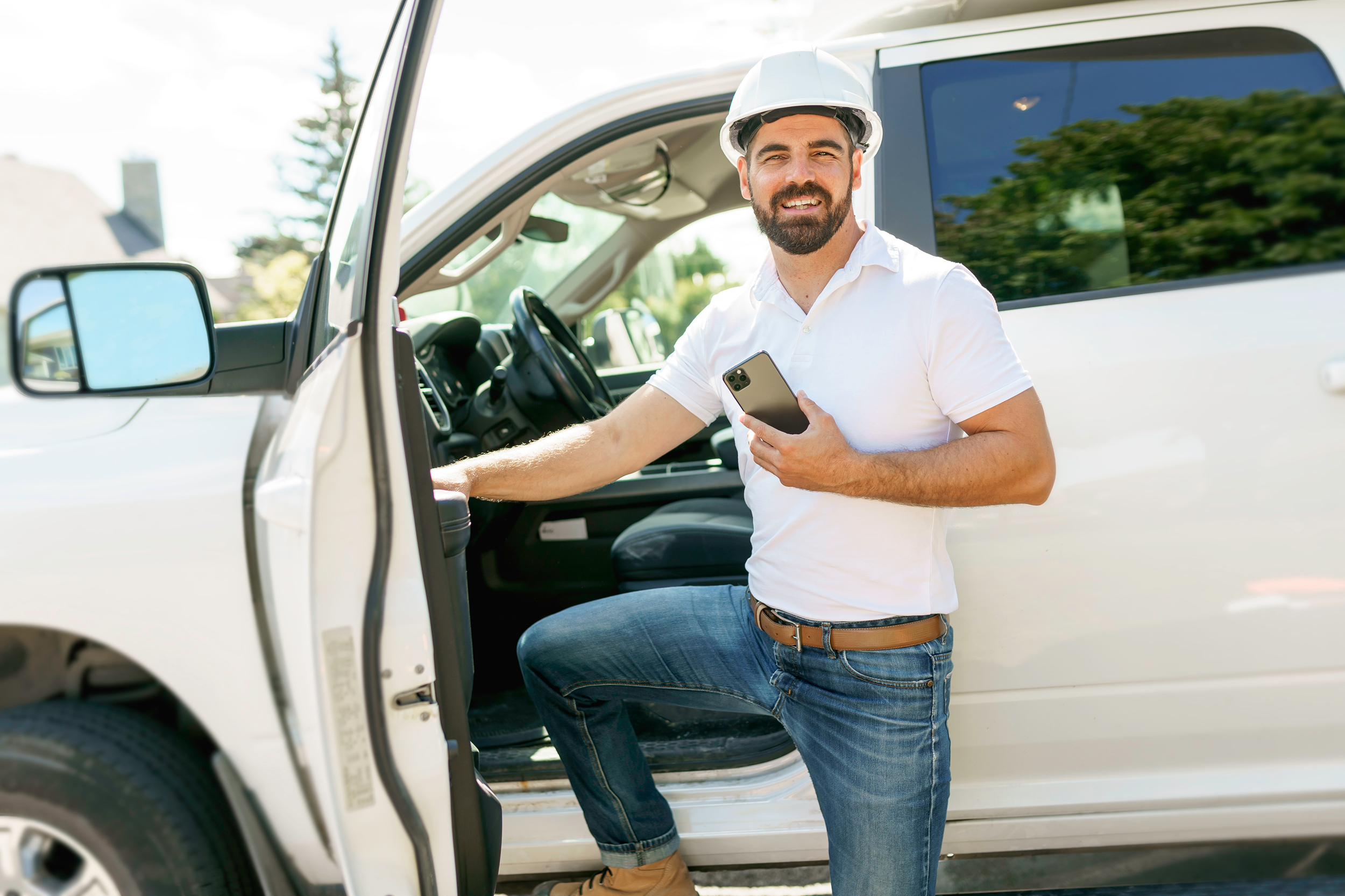 Man holding his phone, wearing a hard hat in front of his work truck