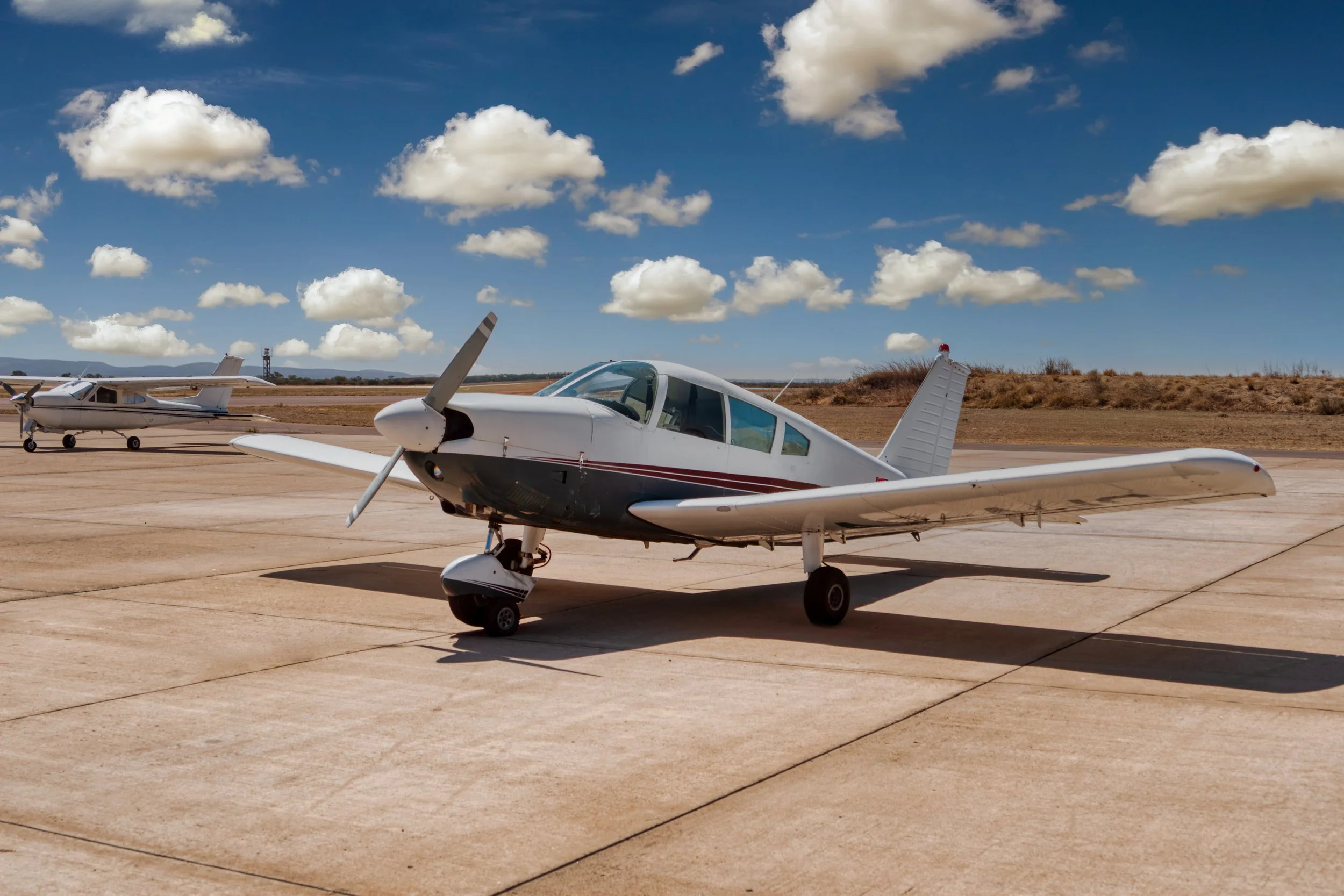 Small private airplane on tarmac with another aircraft in the background, under a blue sky with white clouds.