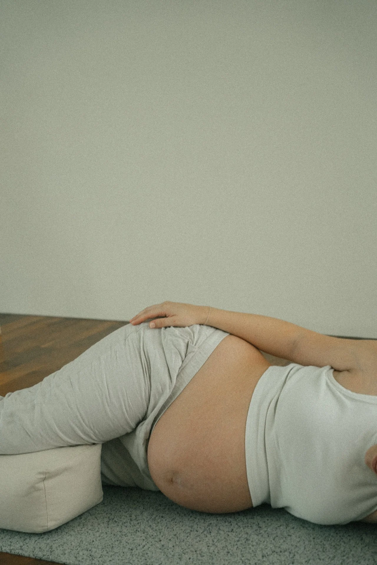 Nahaufnahme einer schwangeren Person, die auf dem Boden liegt und ihren Bauch hält, in einem weißen T-Shirt und grauen Hosen, vor einer neutralen Wand.