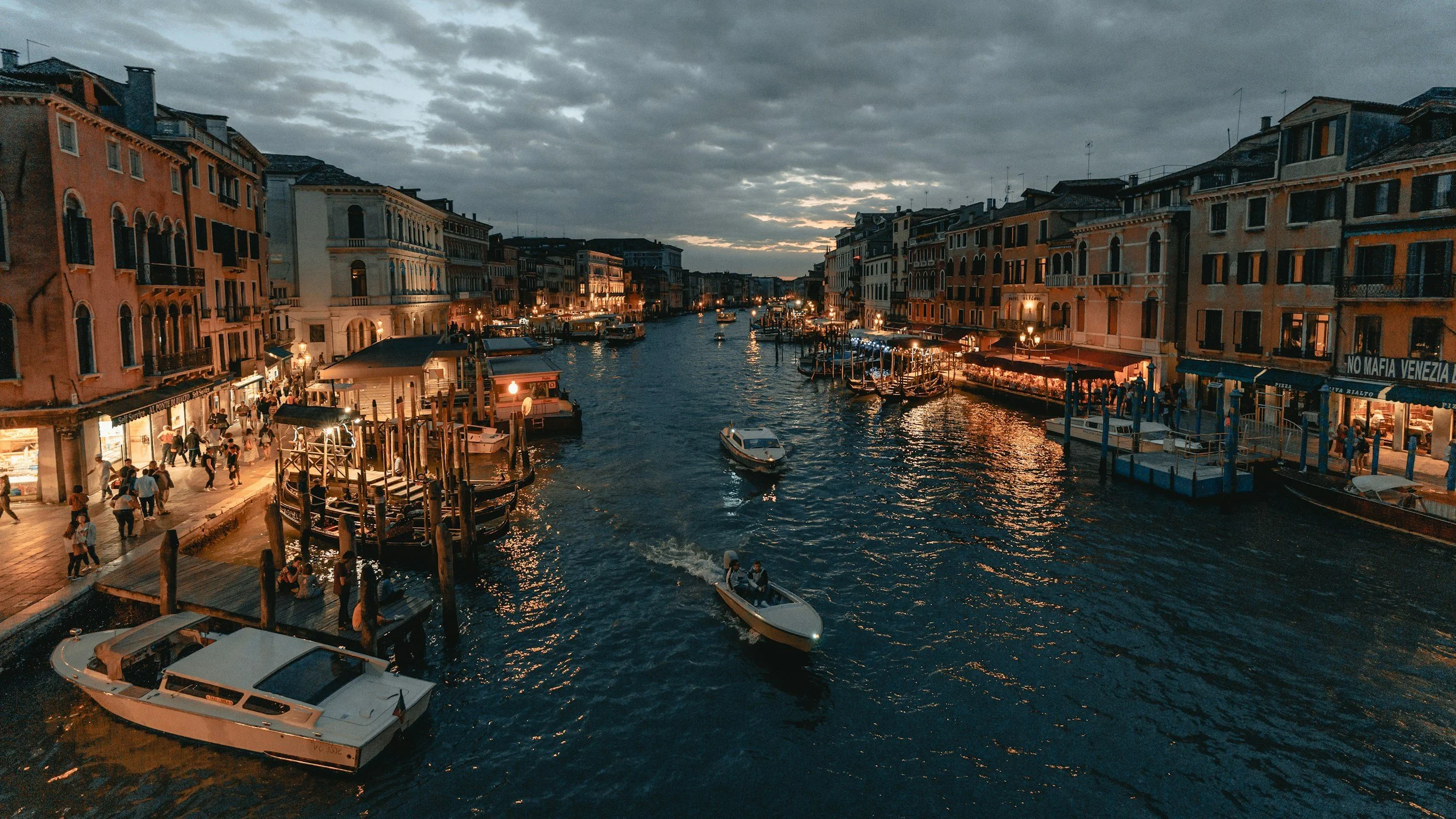 Venetian canal at dusk with boats on the water, adjacent buildings illuminated, people walking on the waterfront.