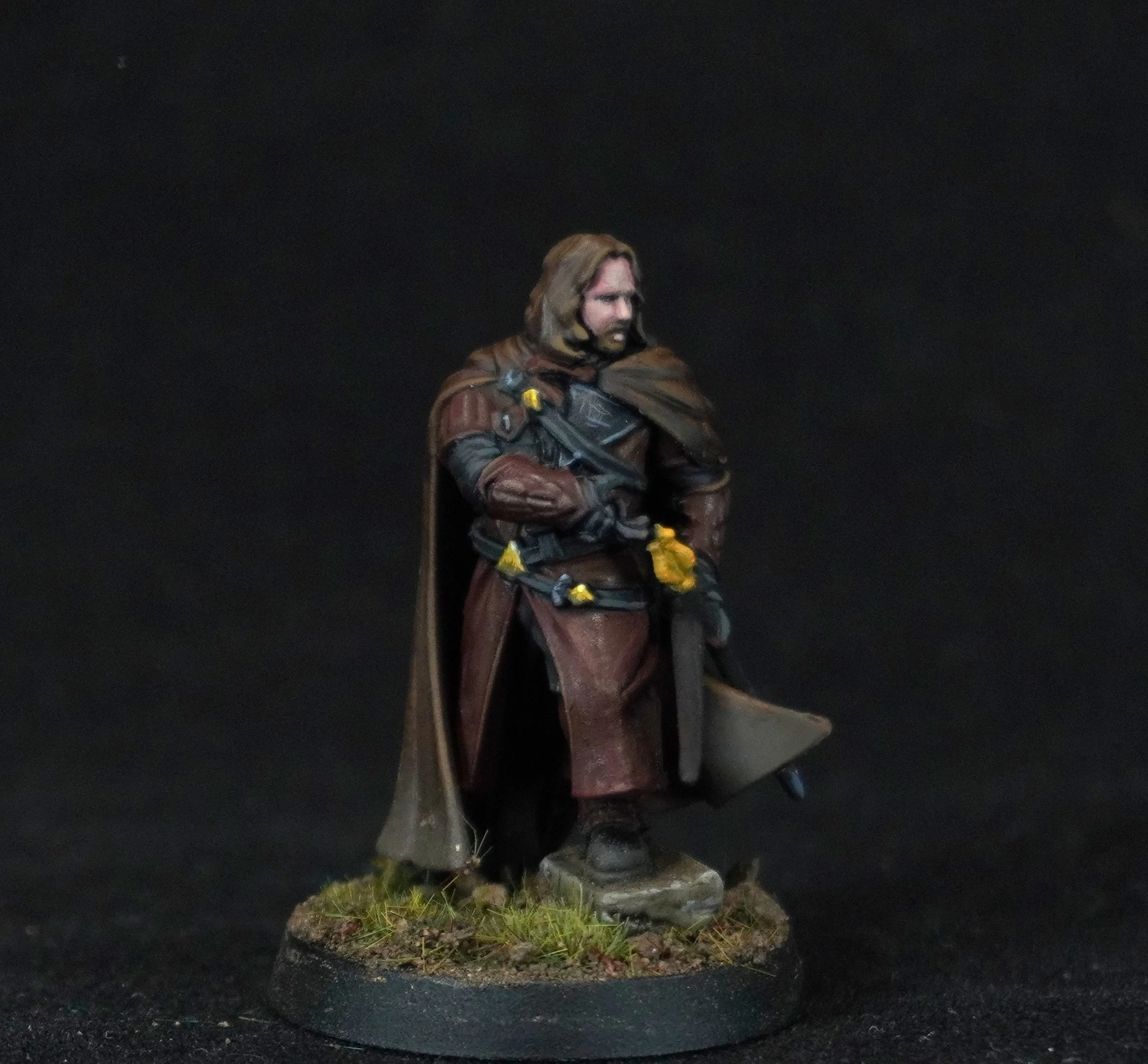 Miniature figurine of a man dressed as a fantasy warrior or rogue, equipped with a sword, wearing a long brown coat, standing on a grass-turf base against a black background.