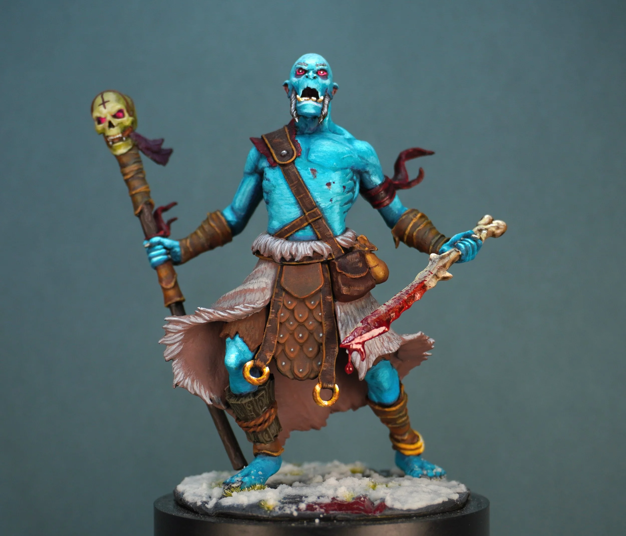 Miniature figure of a blue-skinned, humanoid creature with red eyes and sharp teeth, holding a bloody knife in one hand and a staff with a skull in the other, dressed in primitive armor and standing on a snowy base.