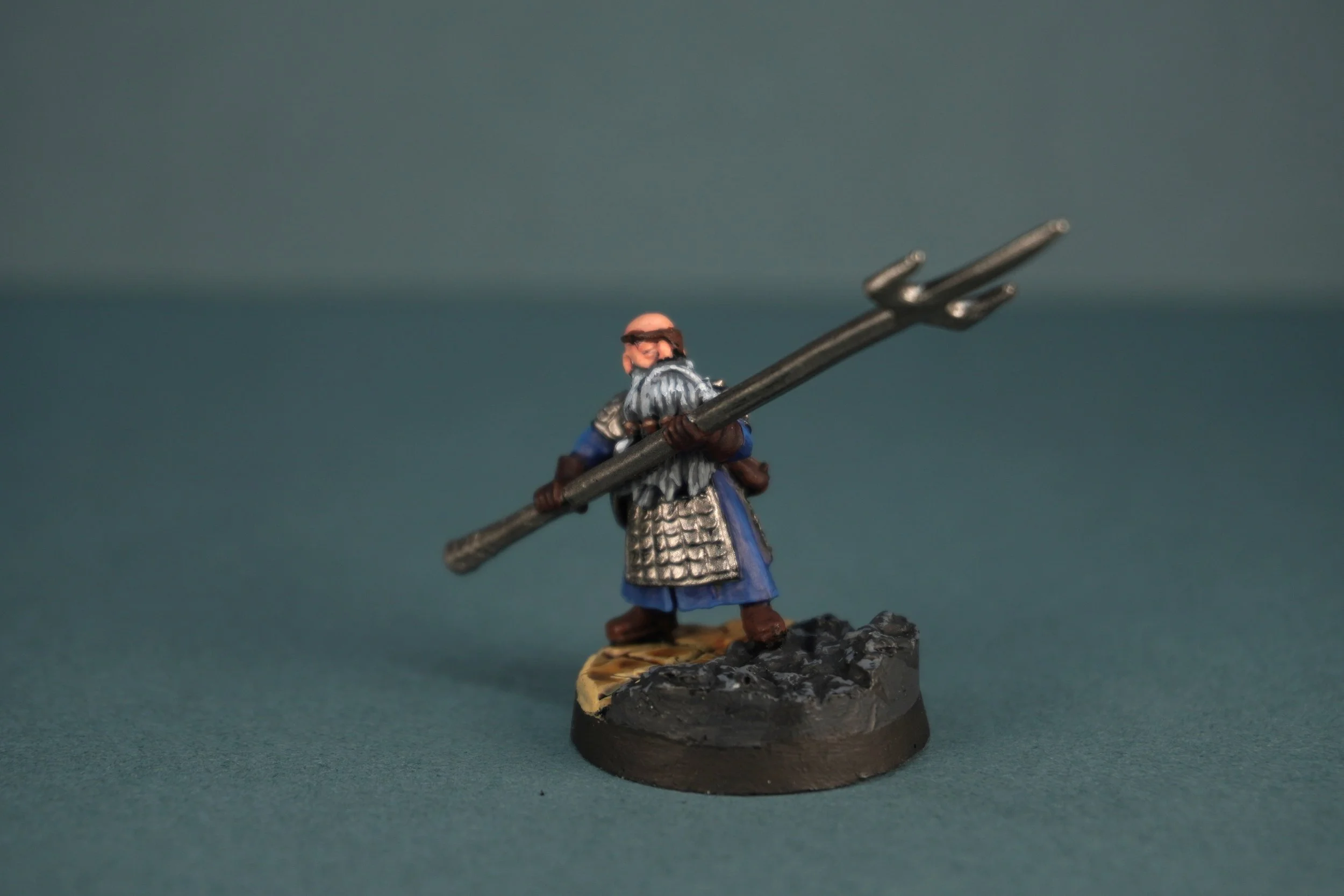 Miniature figure of a dwarf warrior with a long beard holding a large spear, wearing metallic armour and a blue tunic, standing on a textured base.