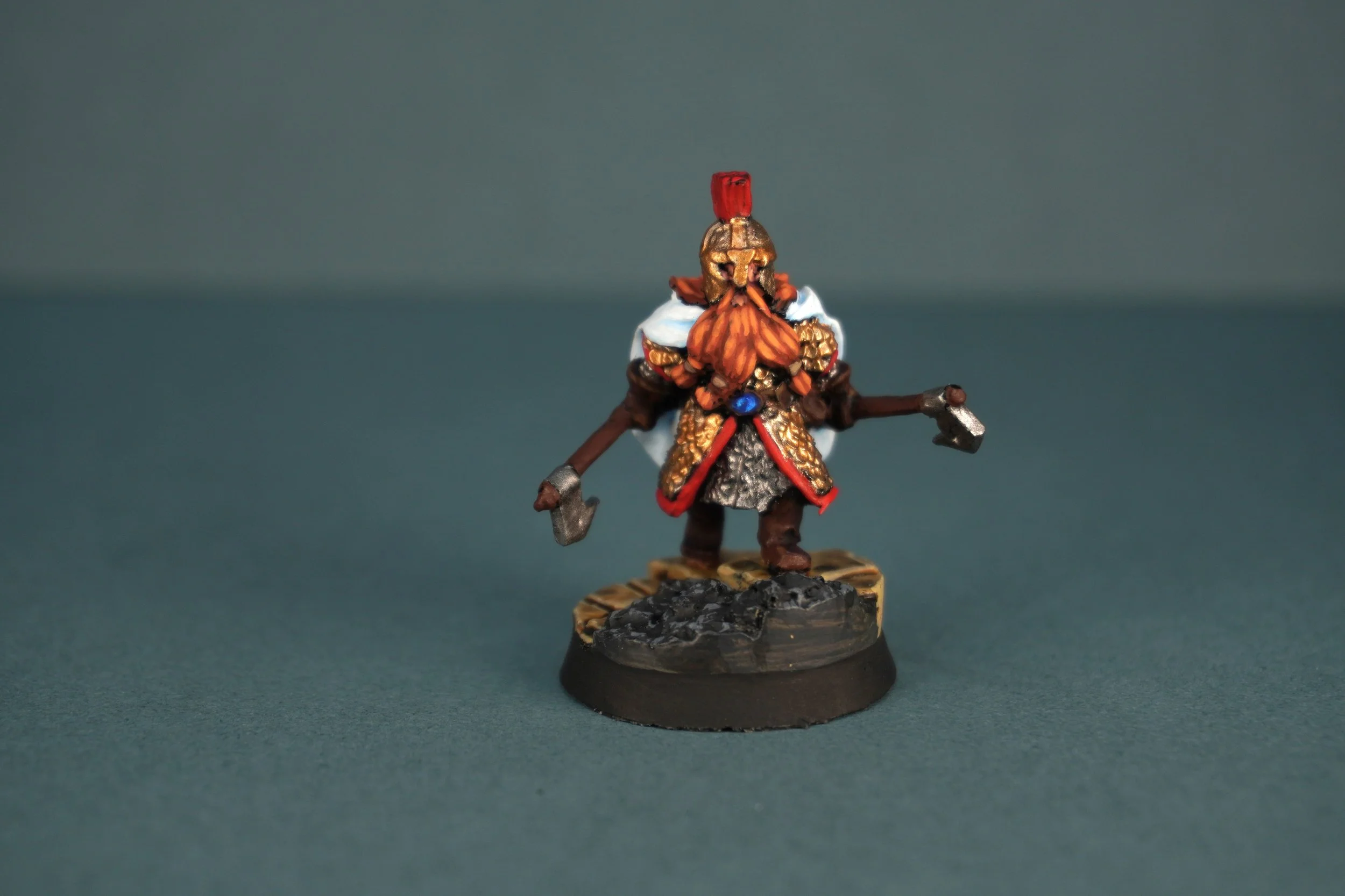 Miniature figurine of a dwarf warrior with a red beard, wearing a helmet and armor, holding two small axes, standing on a rocky base.