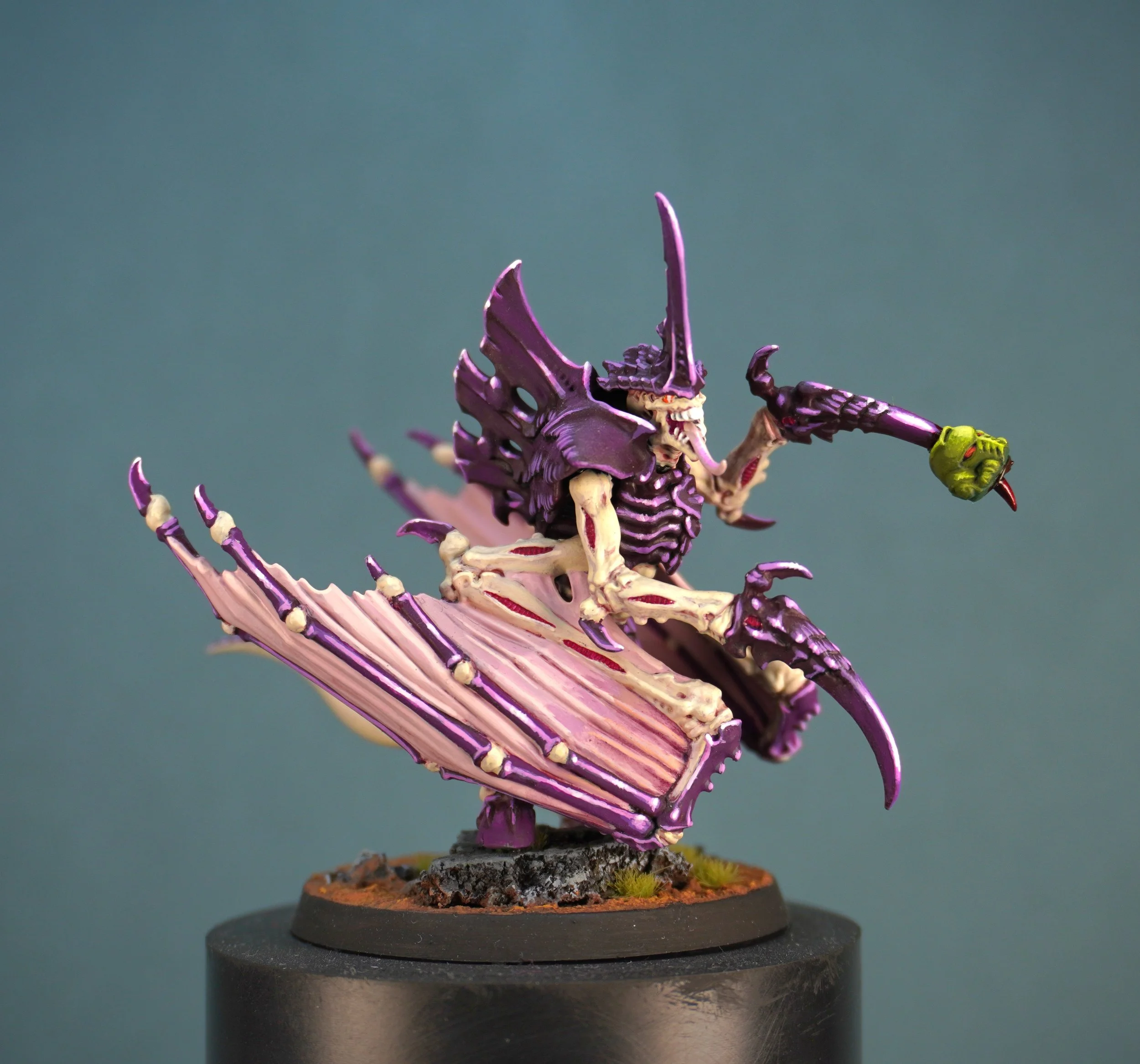 A detailed painted miniature of a purple Tyranid with pink wings, and a salamanders head impaled on his talon. mounted on a round base.