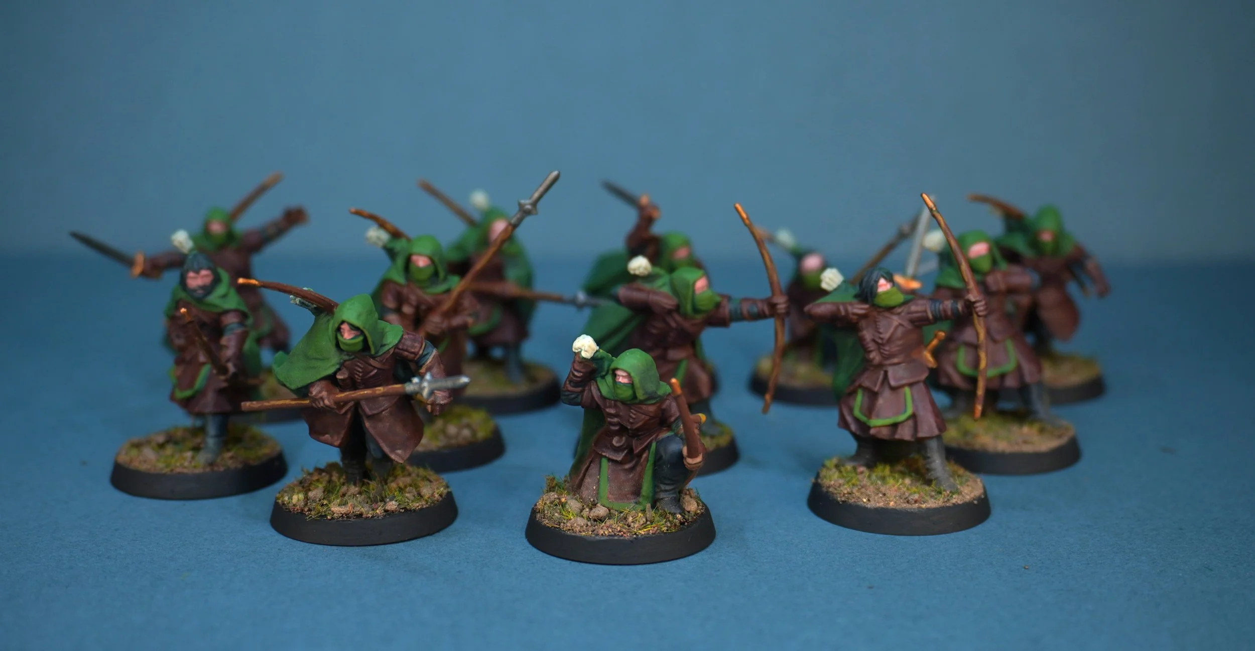 Miniature fantasy soldiers with green cloaks, brown robes, and bows, posed on round bases with grassy textures, set against a blue background.