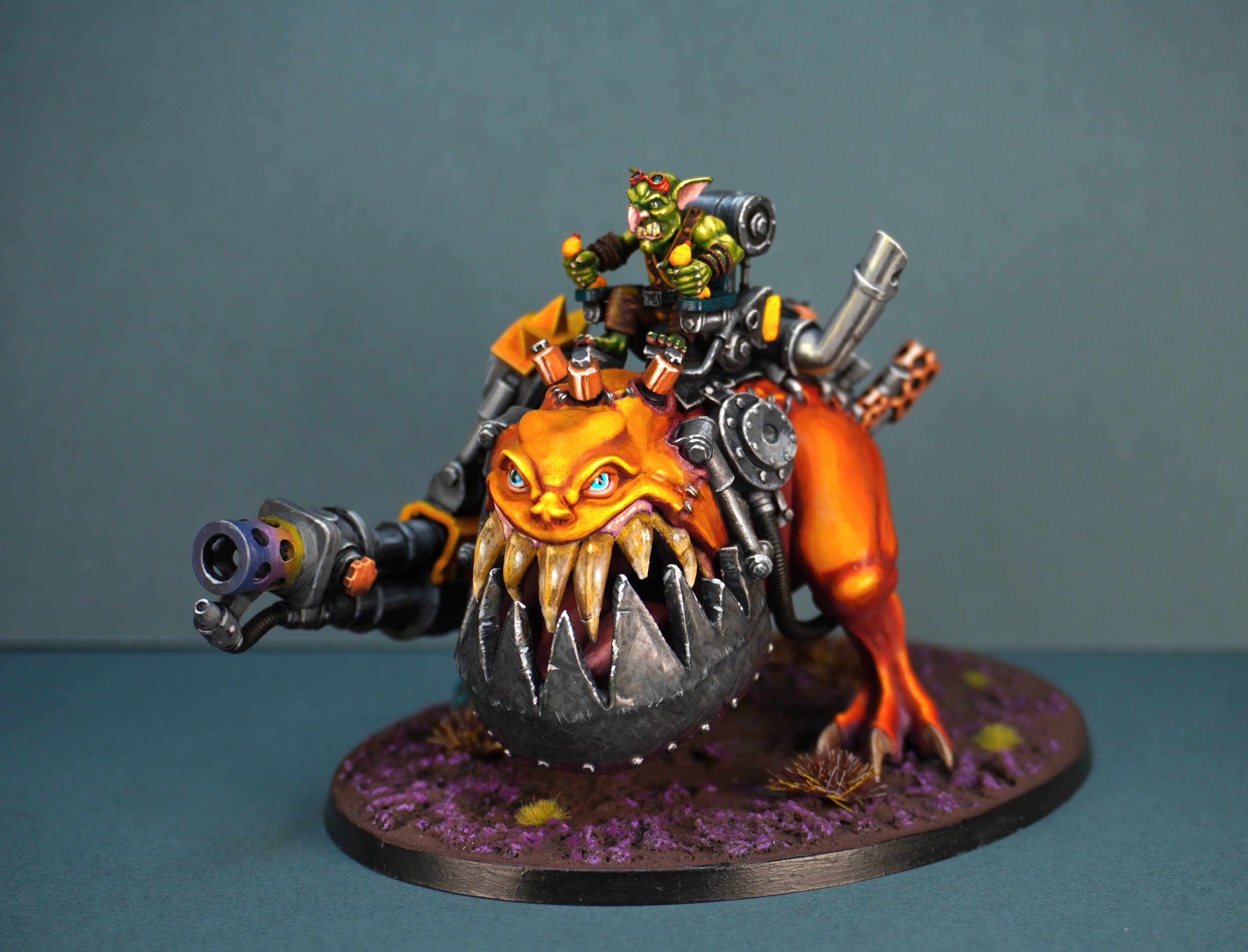 A detailed miniature model of a fantasy creature with a goblin riding a large, fierce-looking beast. The creature has a green face, pointed ears, and wears goggles, while the beast has a large head with sharp teeth, glowing blue eyes, and an orange body. The beast is equipped with mechanical parts and a large gun on its front, and the model is mounted on a textured base with purple and brown ground.