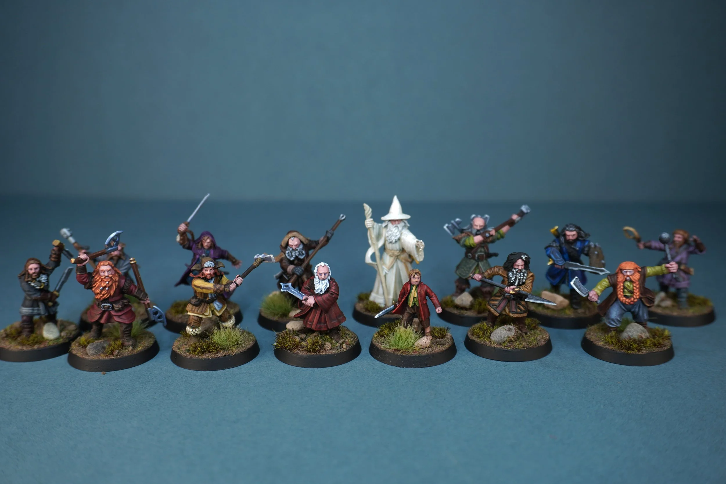Miniature figurines of Lord of the rings characters, including dwarves, Gandalf, and warriors, arranged in a semi-circle on a flat surface with a simple blue background.
