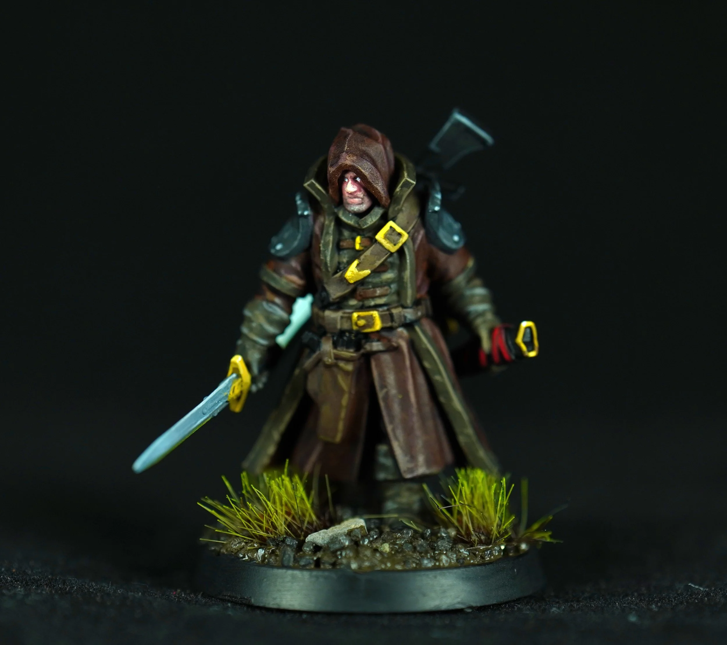 Miniature figure of a adventurer with a hooded cloak, holding a sword, standing on a rocky base with grass, against a black background.