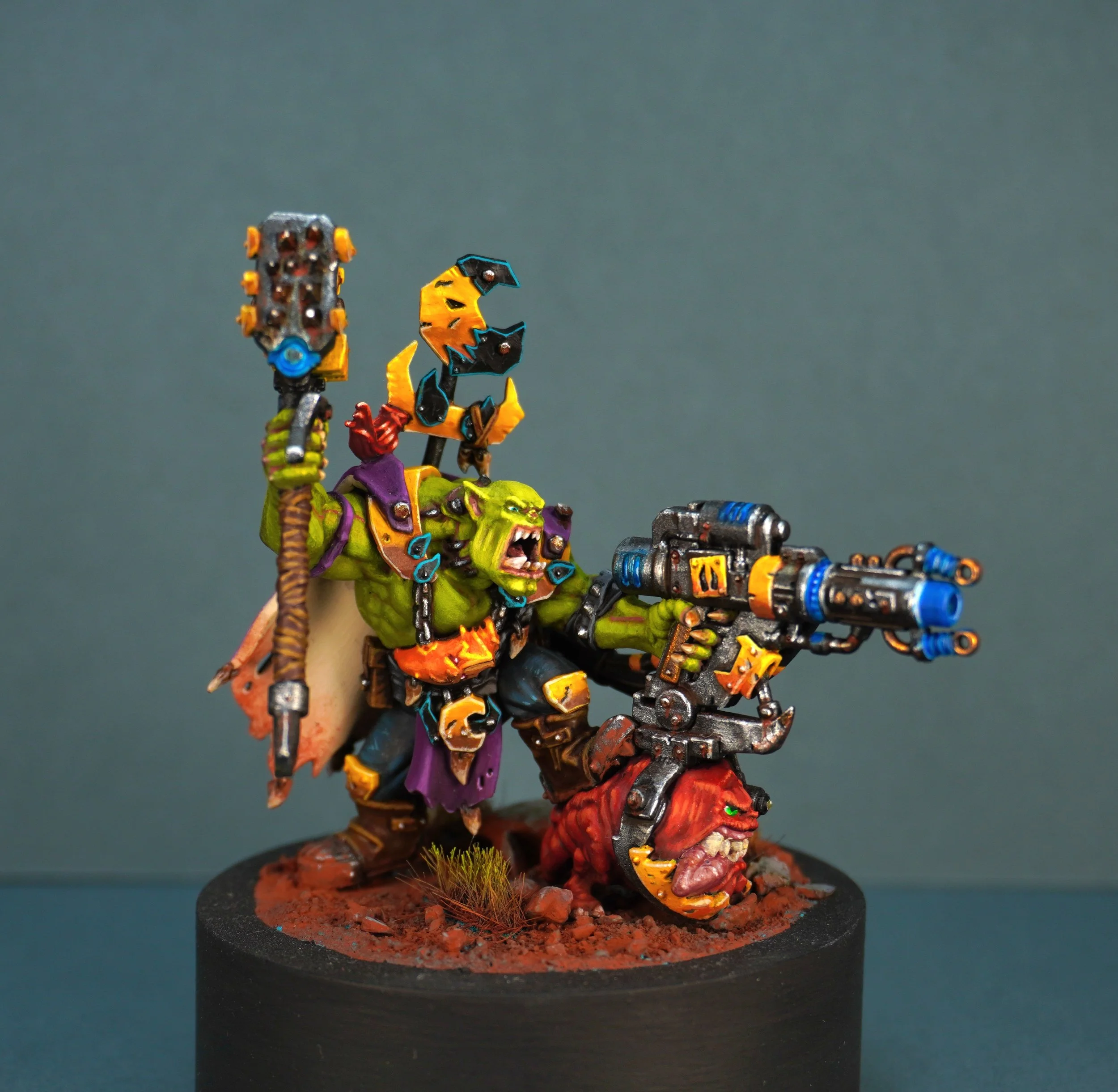A detailed painted miniature of Ufthak Blackhawk wielding a large gun, holding a staff topped with a spiked weapon, wearing yellow armour and a cape, and standing on a textured base with red soil and small grass tufts.