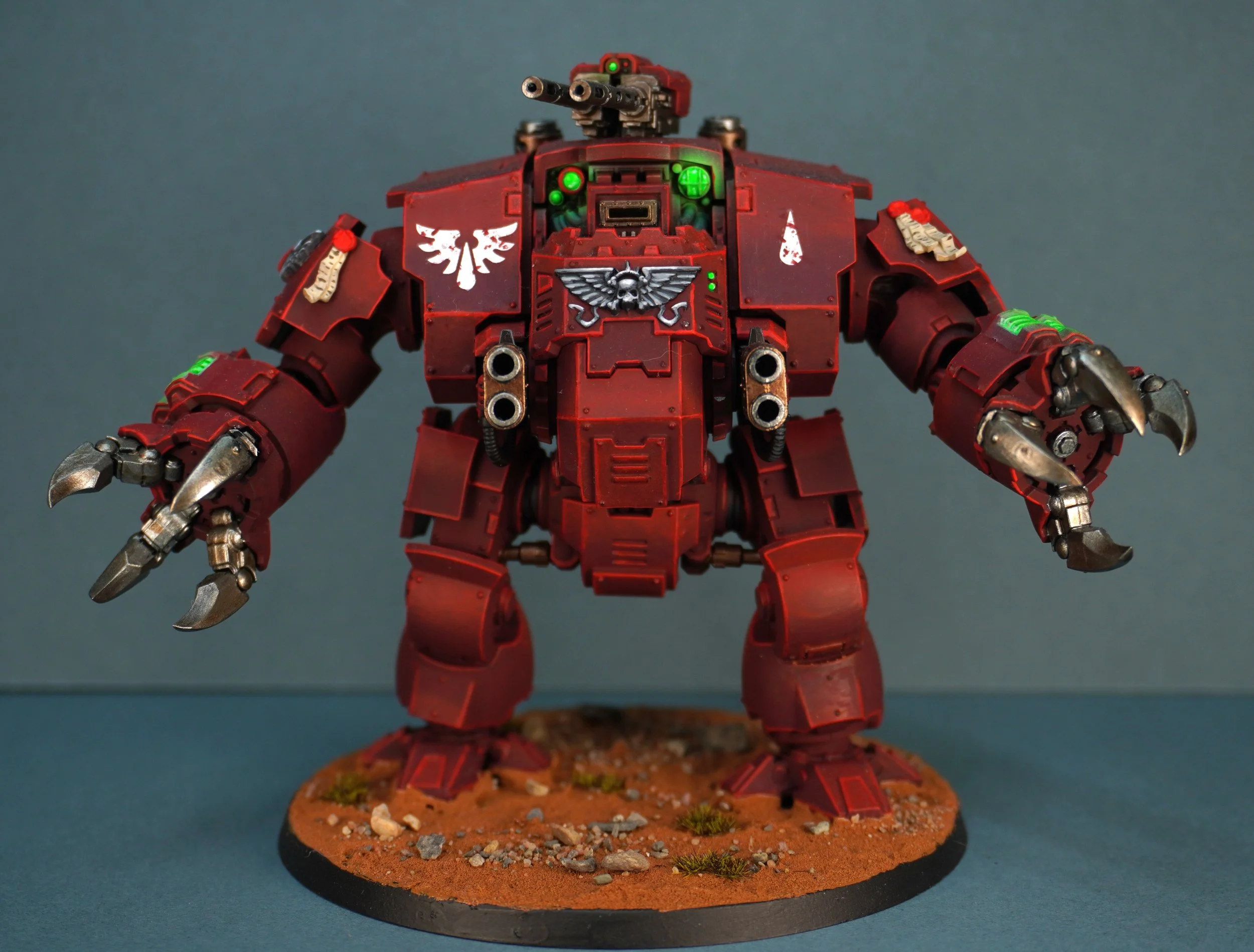 A detailed red miniature model of a battlesuit with mechanical claws, weapons, and symbols, mounted on a textured base resembling a terrain with small rocks and grass.