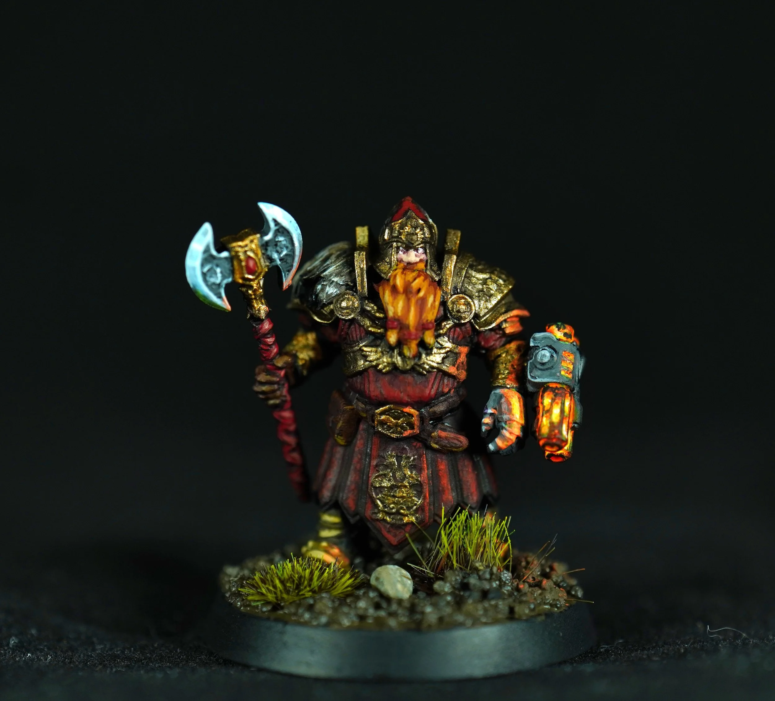 A miniature figurine of a fantasy dwarf warrior, dressed in detailed armor with gold accents, holding an ornate axe in one hand and a small device in the other, standing on a textured base with grass and small rocks, against a black background.