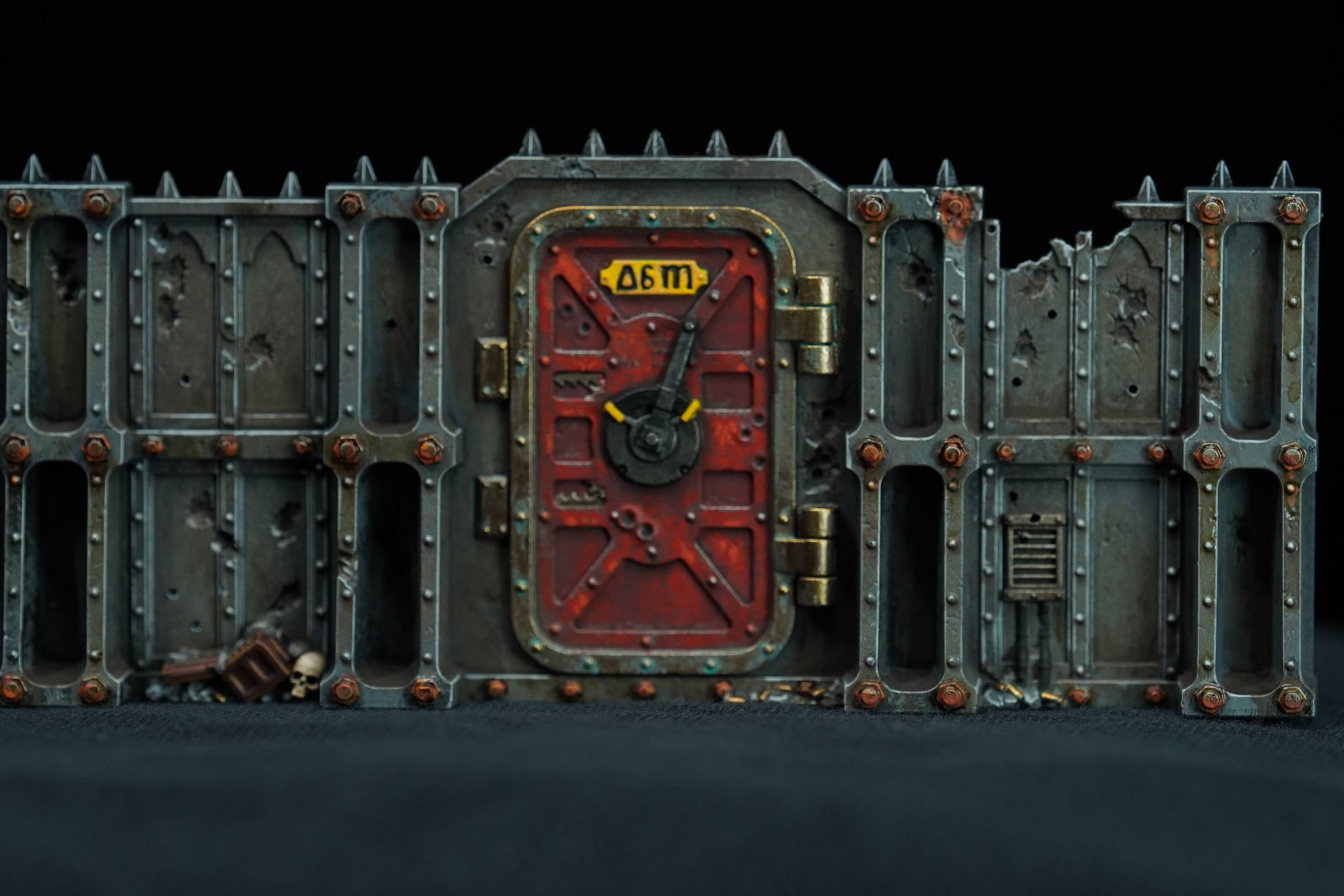 A detailed Terrain piece of a weathered, industrial-style fortress or bunker with concrete walls, Rusty bolts, and a central red door featuring a black wheel and yellow markings, placed on a dark surface with a black background.