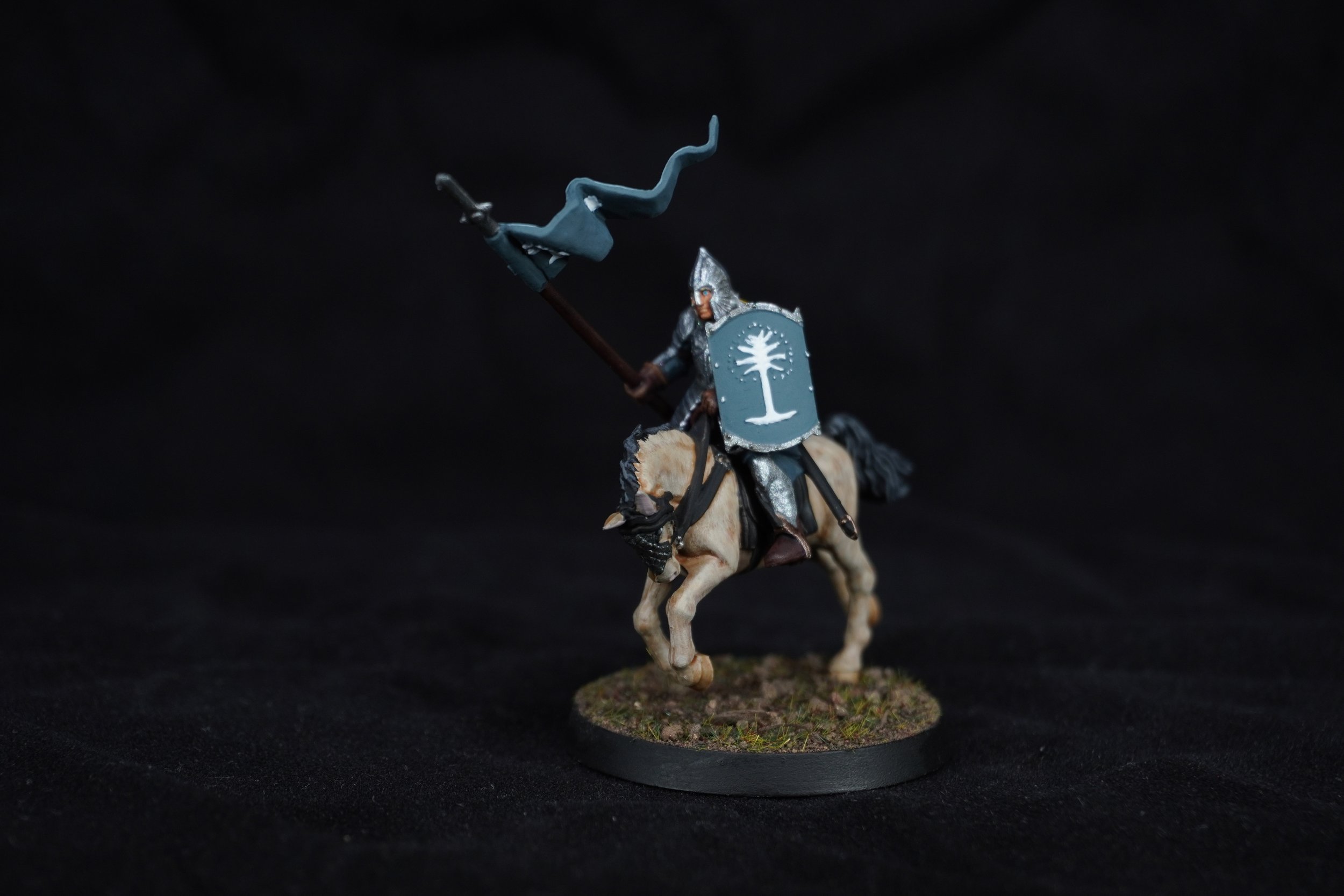 Miniature knight figurine riding a horse, holding a spear and shield, on a round textured base, against a black background.