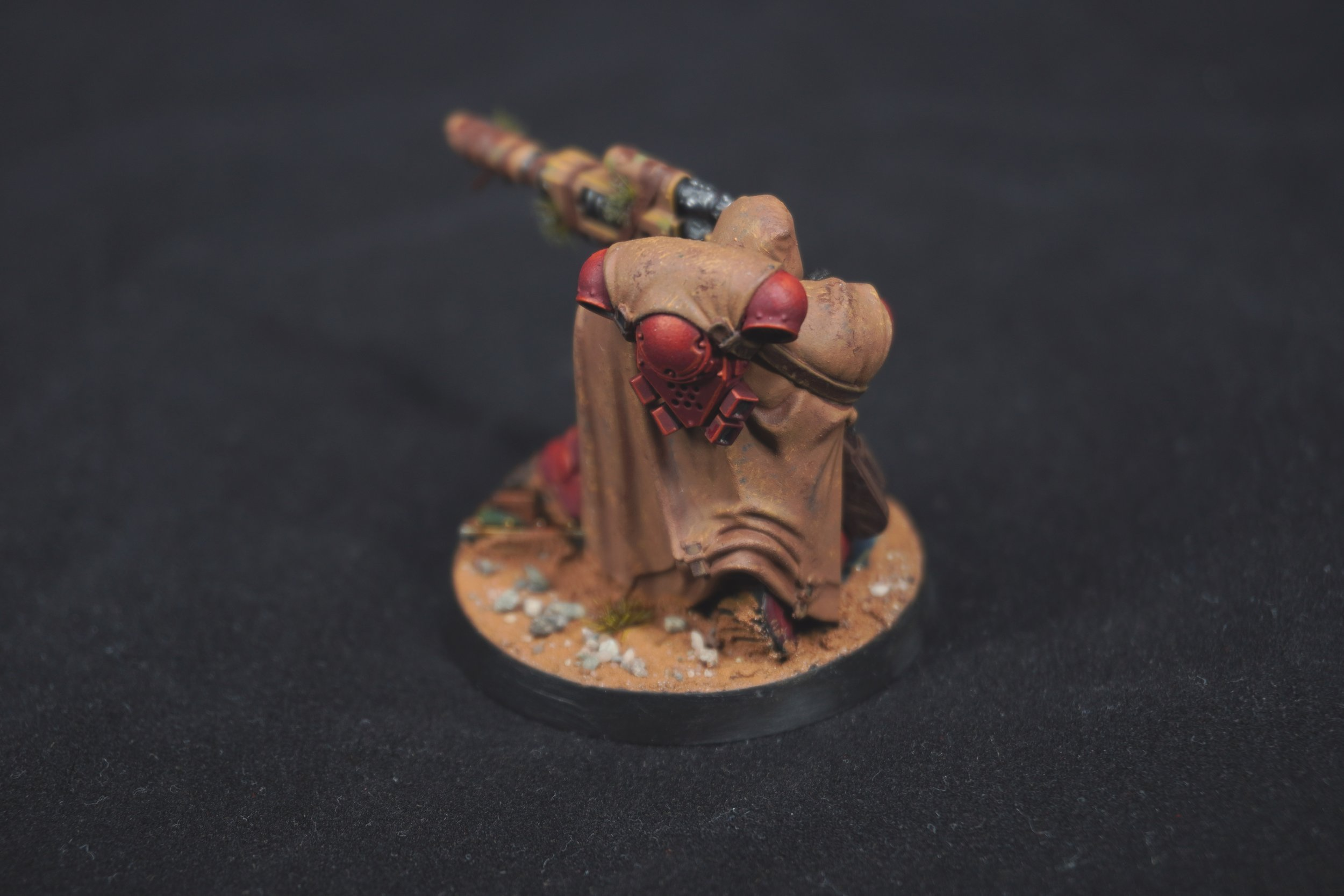 The back side of a painted miniature of a Blood Angel Eliminator, kneeling and aiming a large sniper rifle, on a textured base with sand, rocks, small plants, against a black background. Painted by Suffolk War Painting Studio