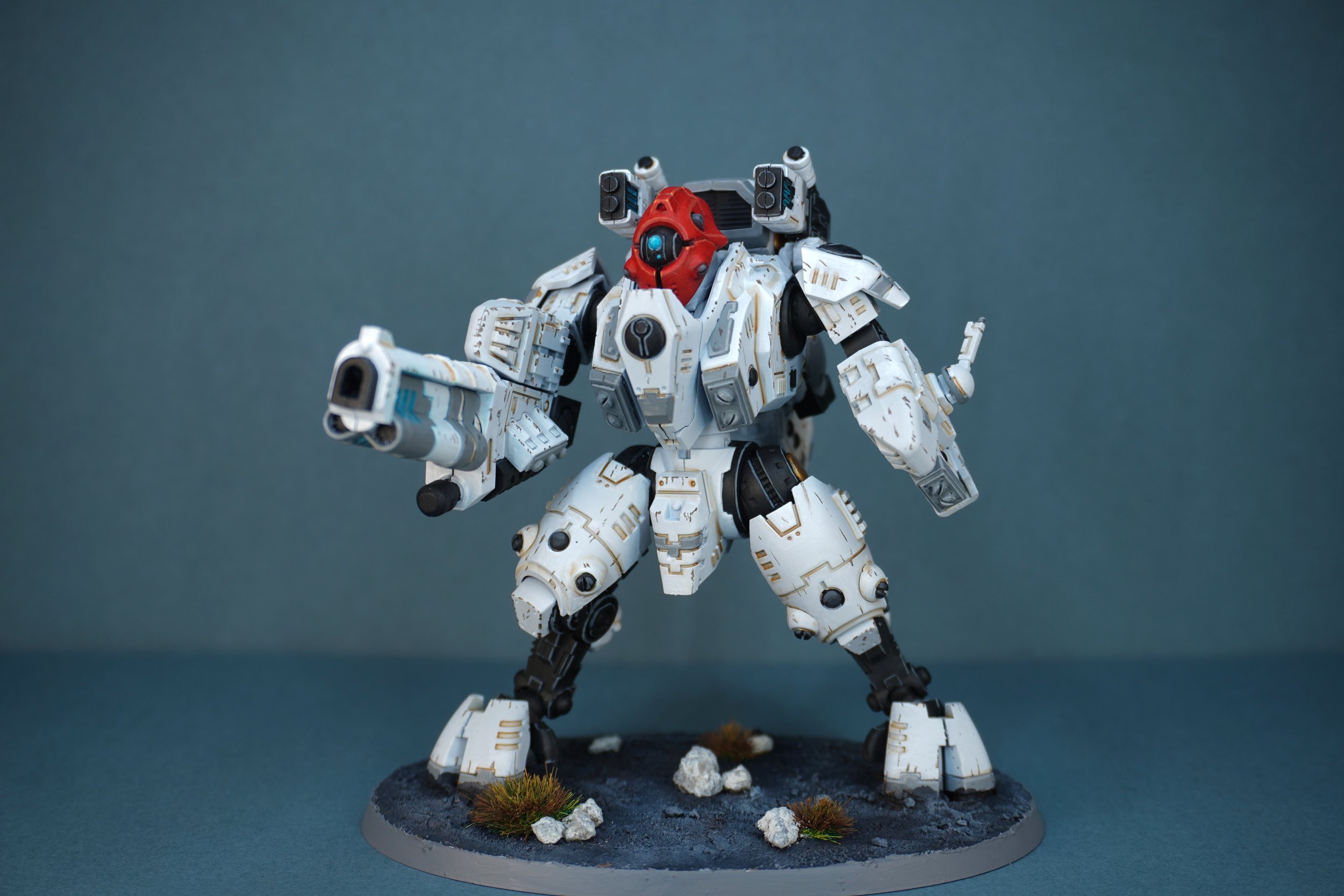 A detailed model of a Tau Ghostkeel from Warhammer 40k in a white and grey armour, black joints, and a red headplate, standing on a circular display base with rocks and grass, against a plain background.