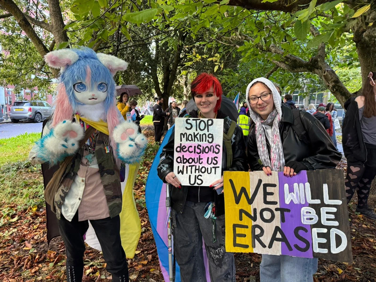 Signs: "Stop making decisions about us without us" and "We Will Not Be Erased" on a non-binary flag