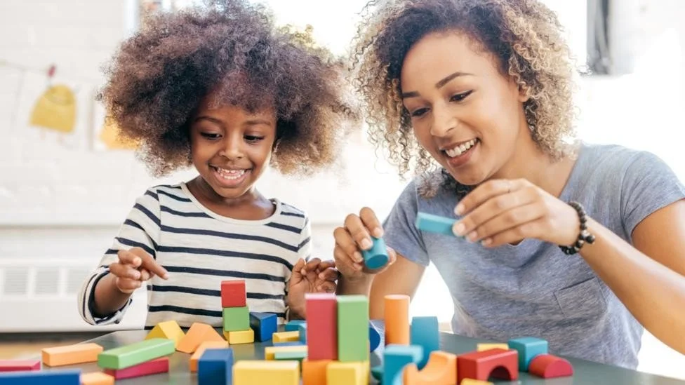 A woman and a young girl playing with colorful wooden blocks at a table, smiling and enjoying their activity.