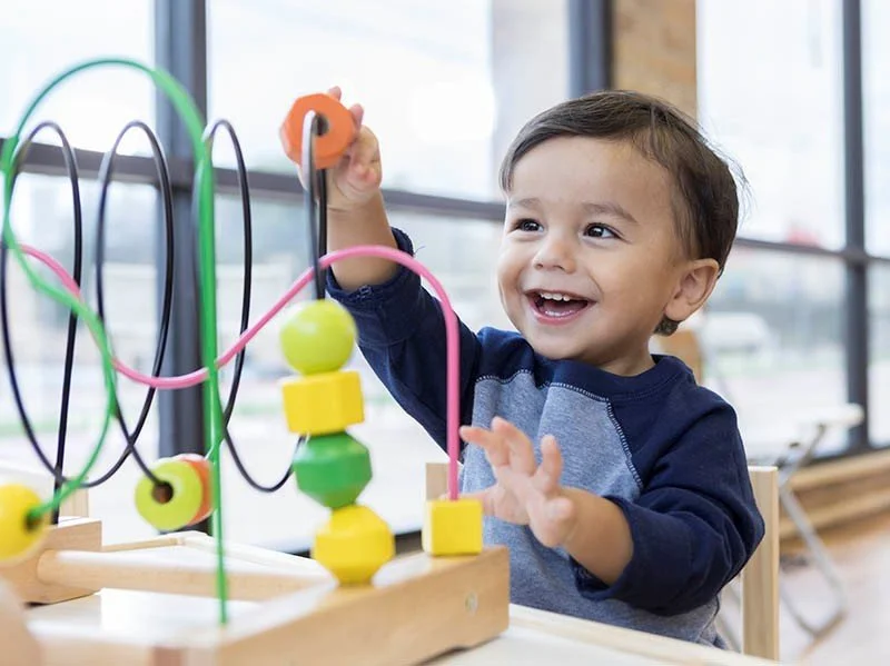 Smiling young boy playing with a colorful wooden bead maze toy in a bright room with large windows.