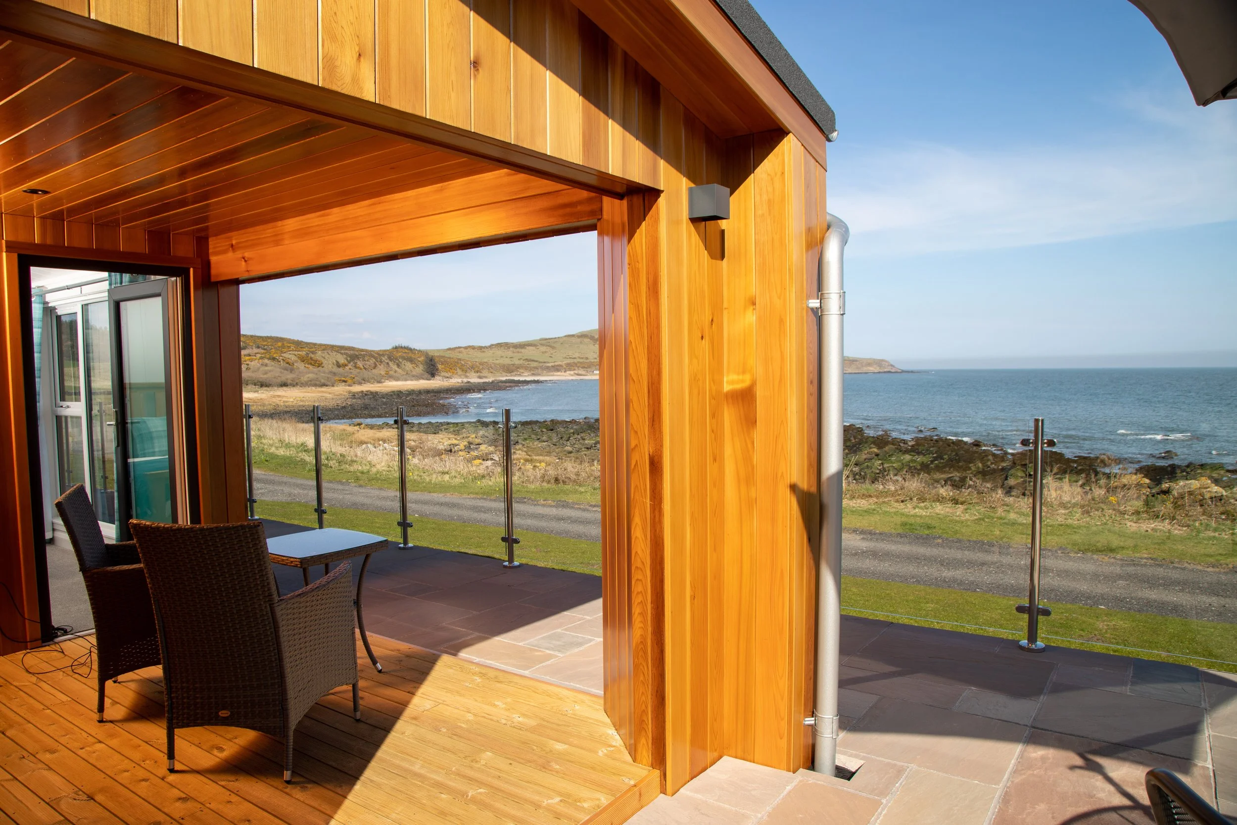 Ocean view from a wooden balcony with patio furniture and a glass railing, overlooking a rocky shoreline and distant hills on a sunny day.