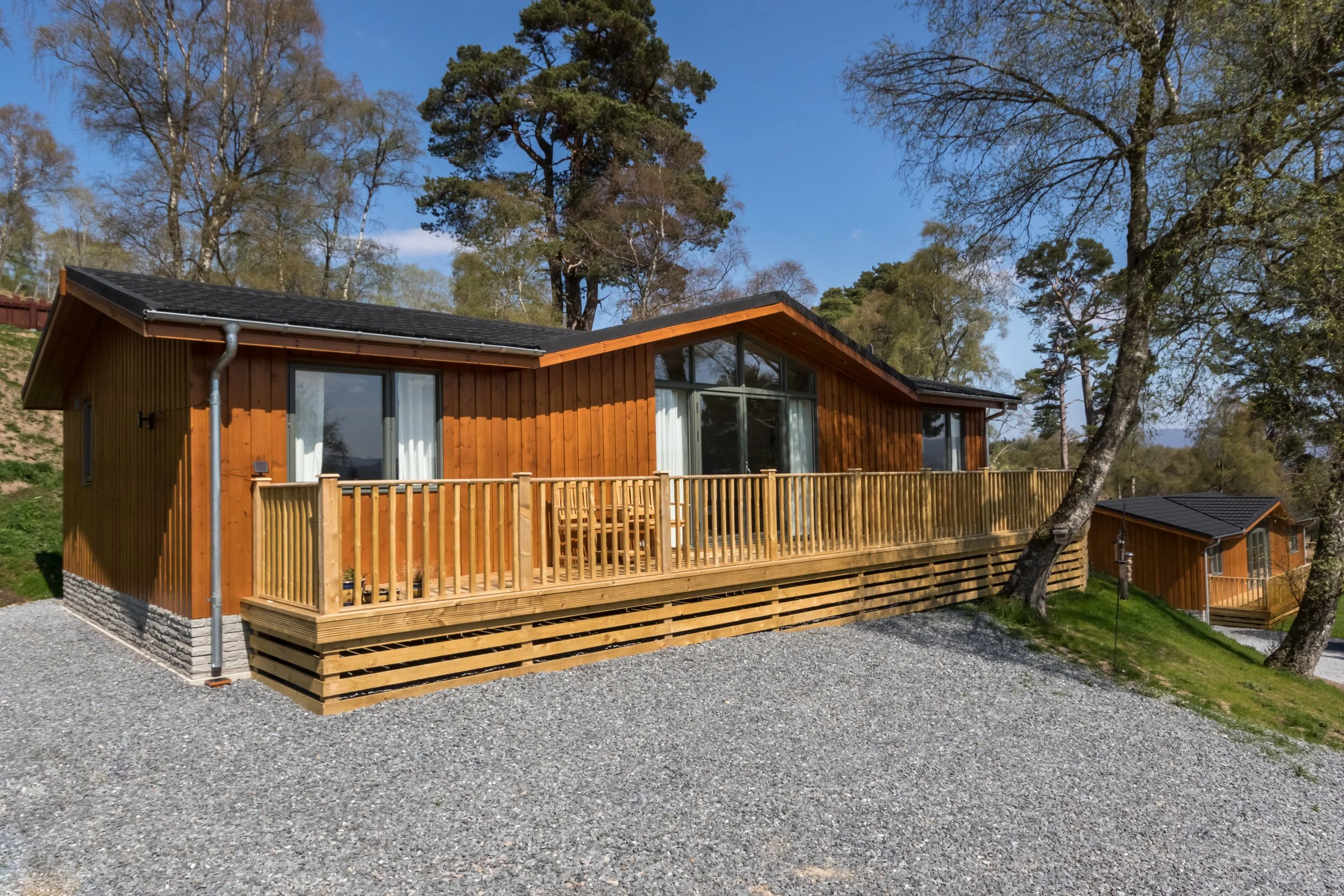 A wooden house with a sloped roof and a large front deck, surrounded by trees and a gravel driveway.