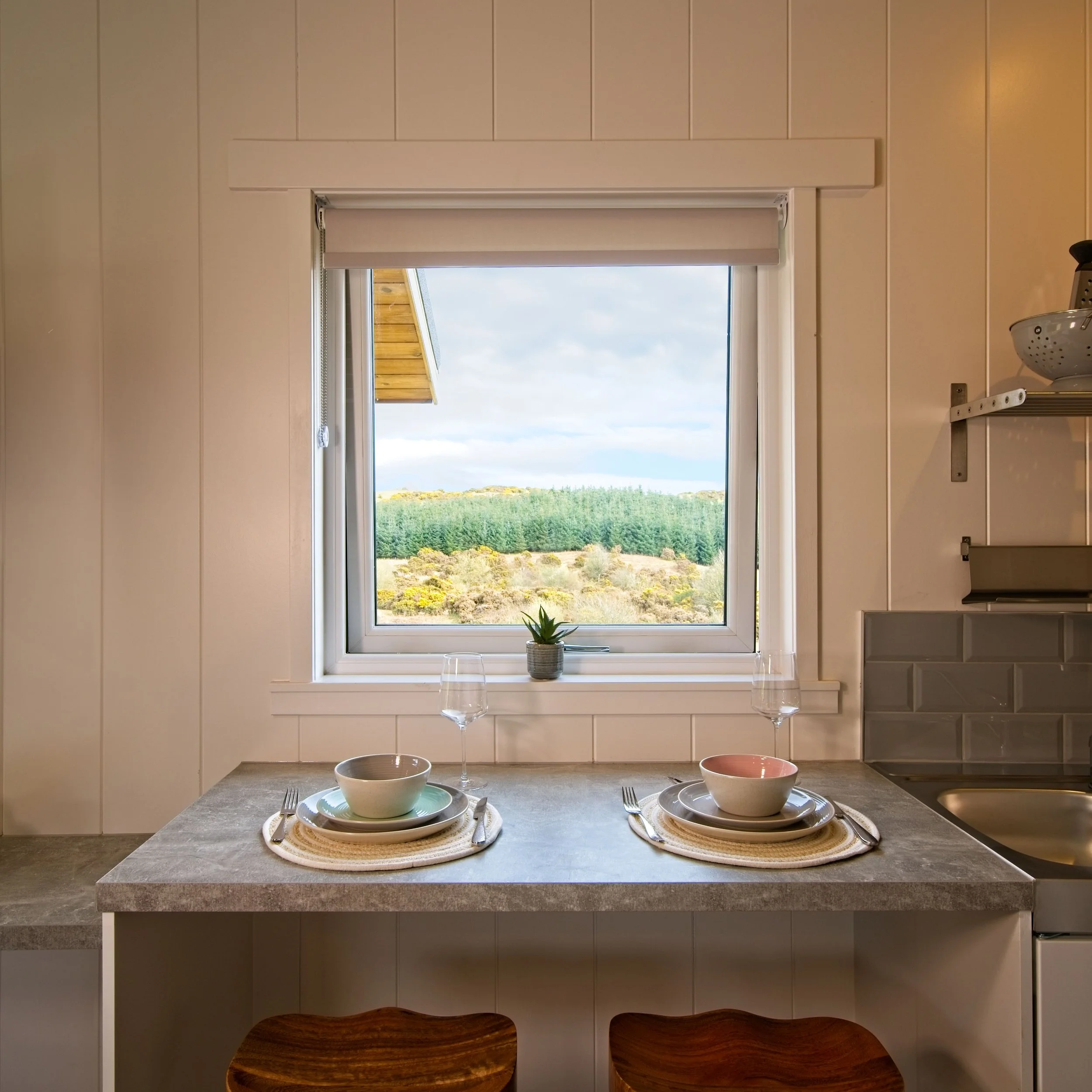 A kitchen counter set for two, with bowls, plates, utensils, and wine glasses, positioned in front of a window showing a landscape with trees and hills.