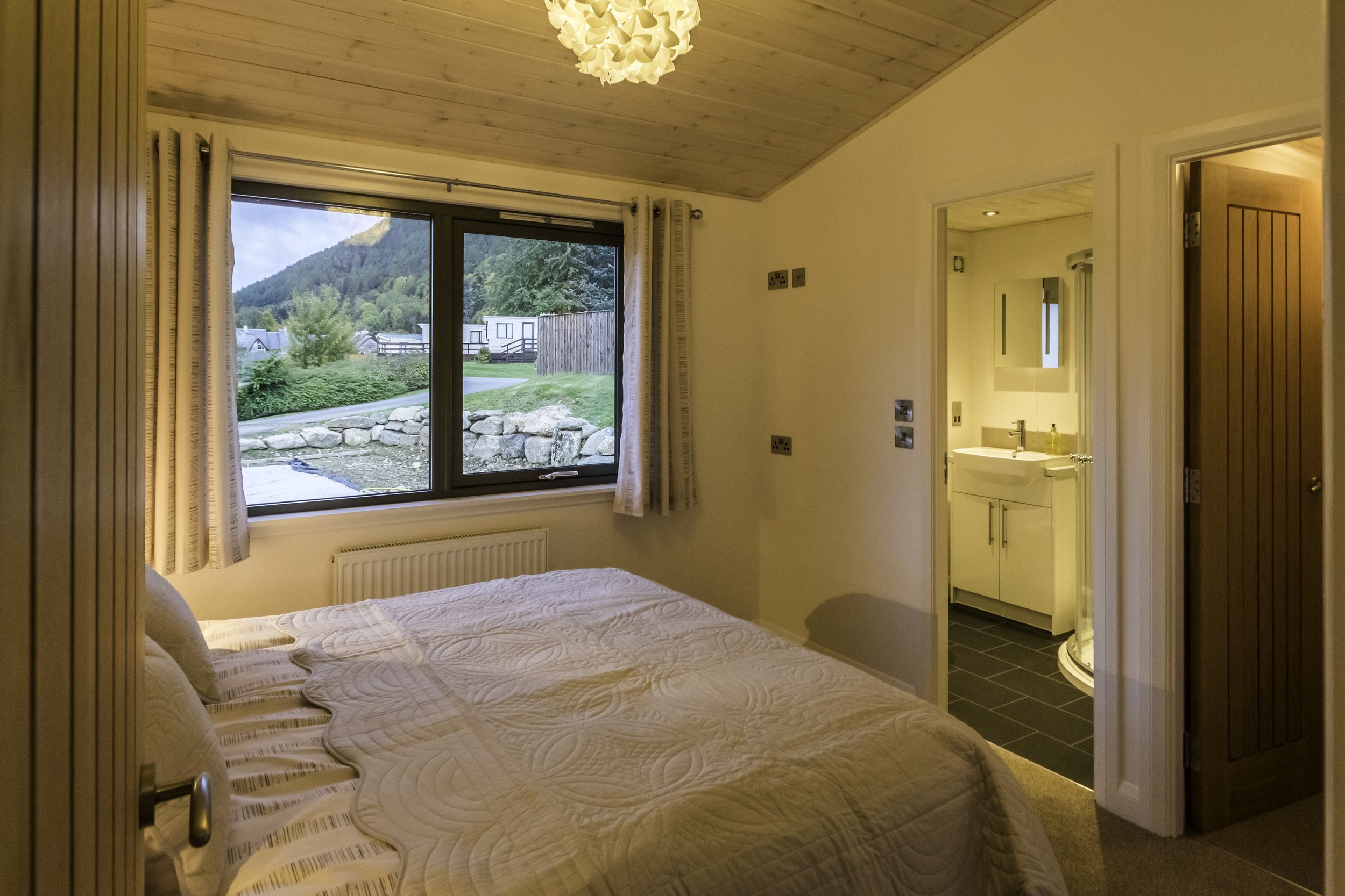 A cozy bedroom with a large window revealing a green landscape and mountains outside. The room has a wooden ceiling, a bed with a quilted cover, and an adjacent bathroom with a sink and mirror.