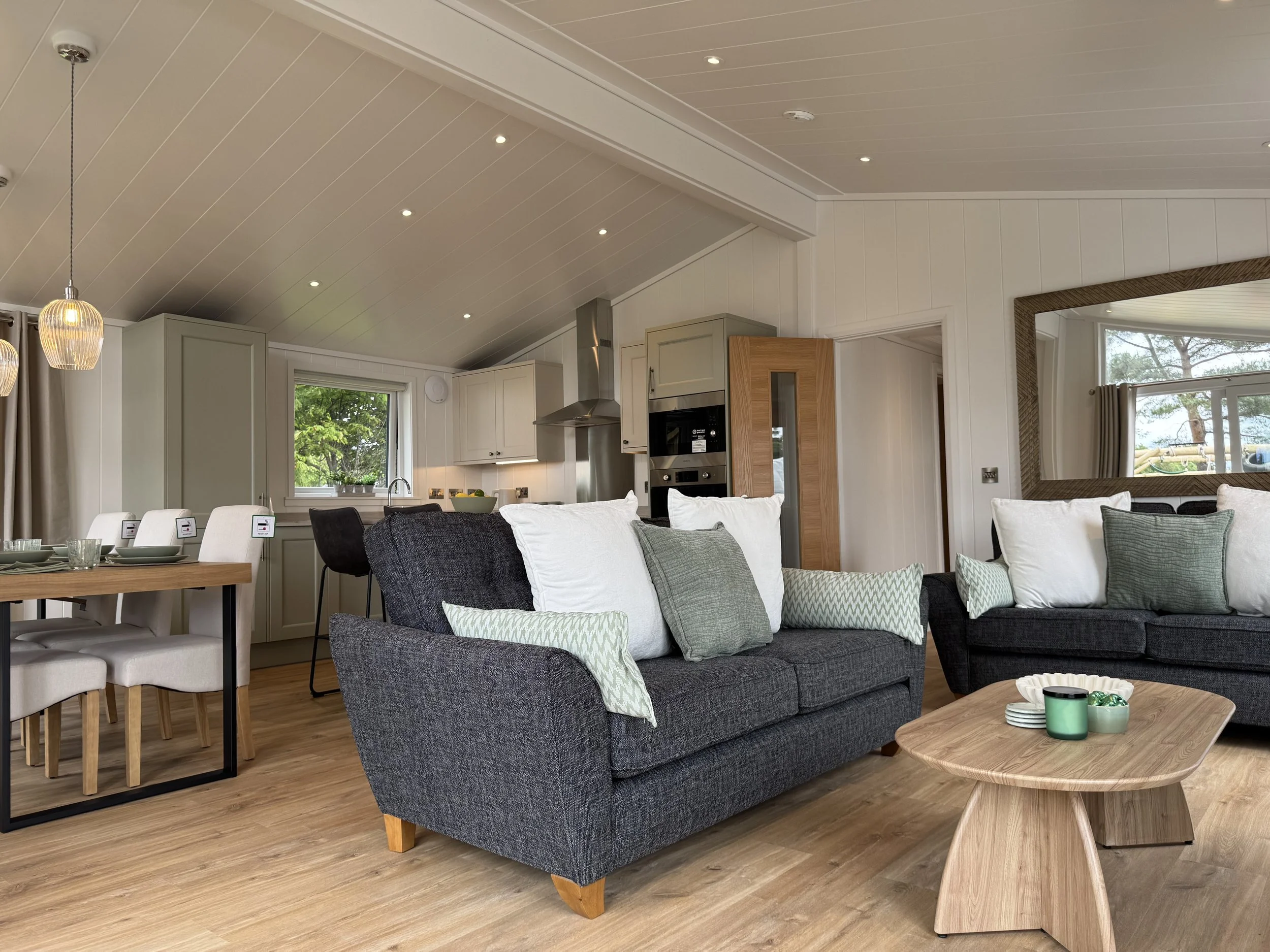 Living room and kitchen in a modern home with wooden floors, dark gray couches with white and green pillows, a wooden coffee table, dining table with white chairs, and a kitchen with white cabinets and stainless steel appliances.
