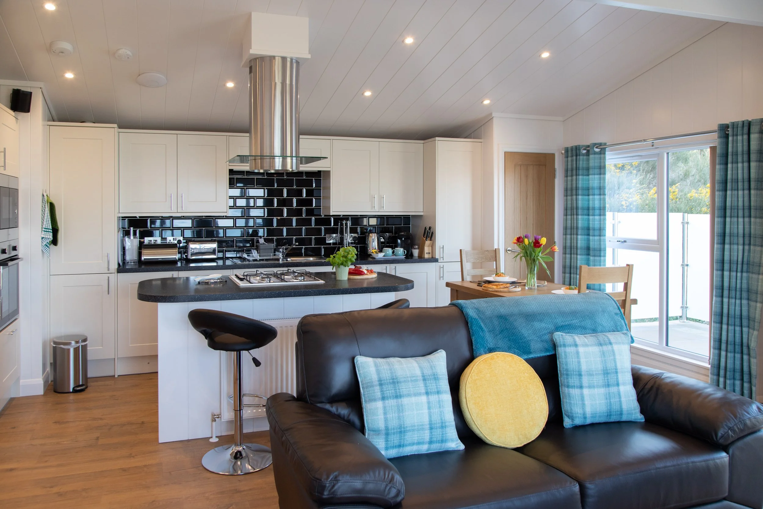 Open-concept kitchen and living room with black leather couch, dining table with flowers, and sliding glass door to outside deck.