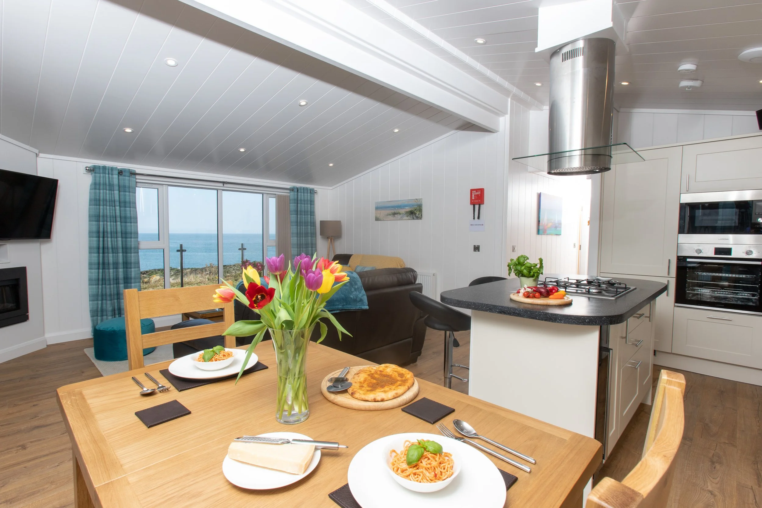 A cozy open-concept living and kitchen area with a view of the ocean through large windows, decorated with colorful flowers, set for a meal featuring pasta and pizza.