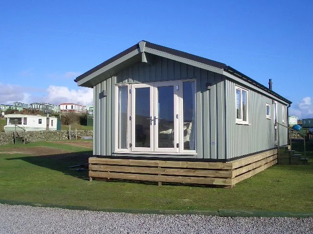 A modern tiny house on a raised wooden foundation with a glass front door, situated on a grassy area under a blue sky.