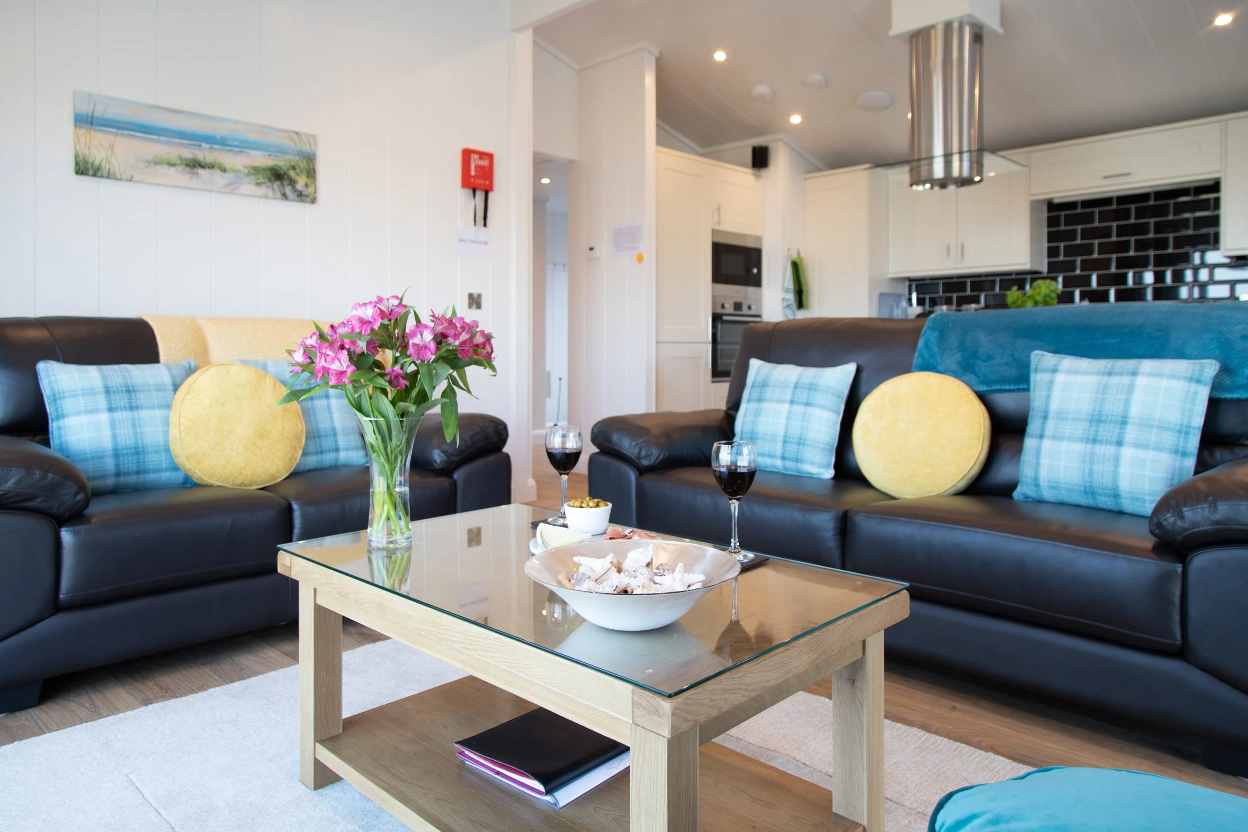 Living room with two black leather sofas decorated with plaid and solid yellow pillows, a glass-top coffee table with a bowl of snacks, glasses of wine, and a vase of pink flowers, kitchen in the background with white cabinets and black tile backspla
