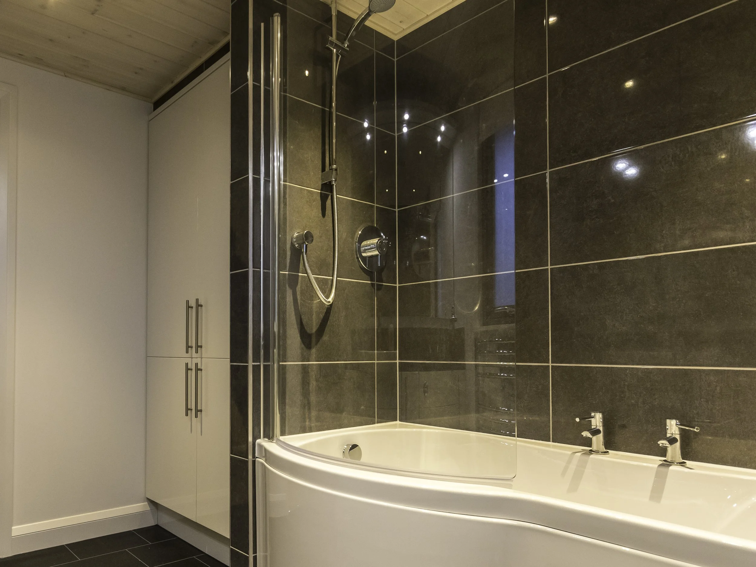 Modern bathroom with a corner shower and bathtub combination, dark tiled walls, a white cabinet with silver handles, and a wooden ceiling.