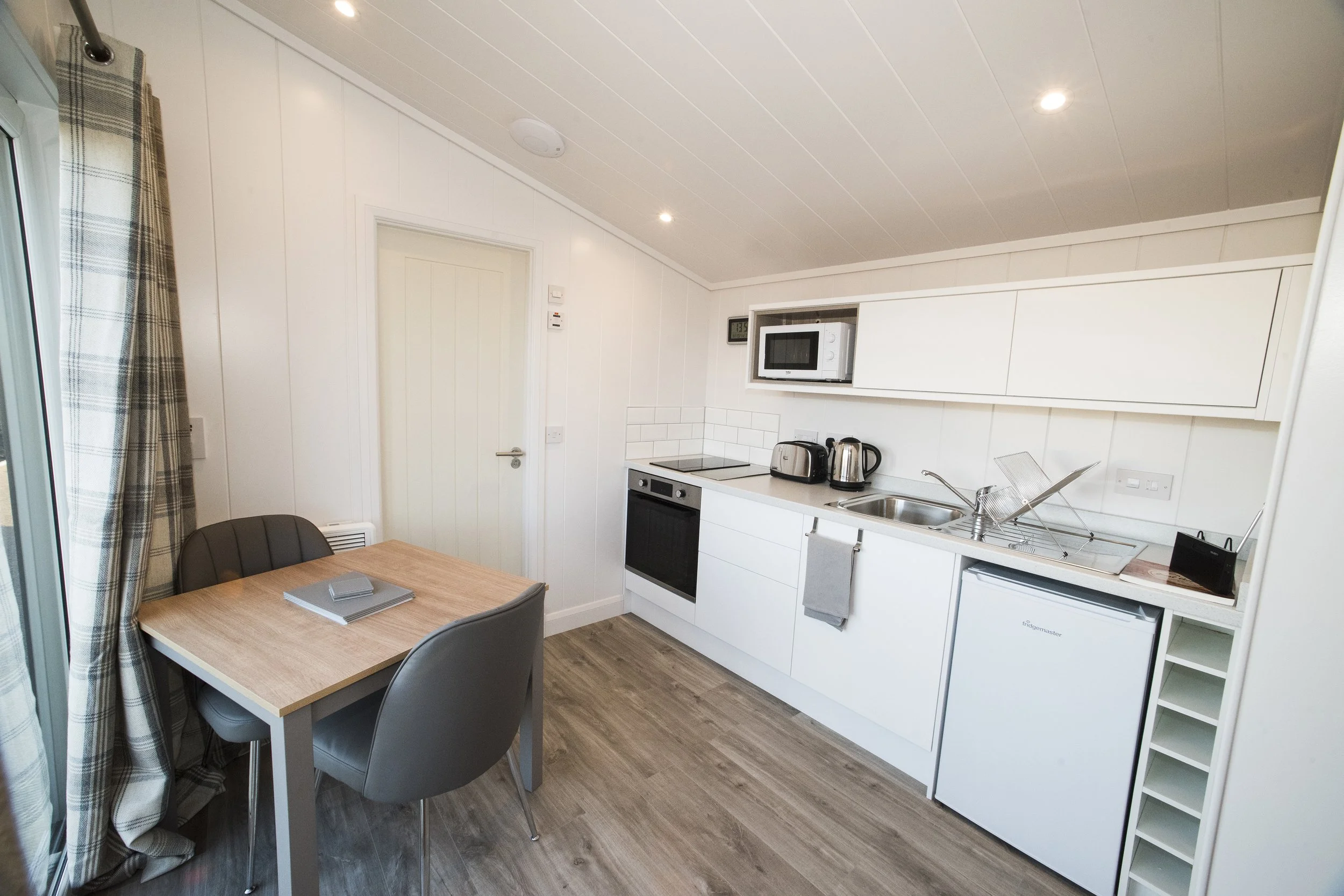 A small kitchen with white cabinets, a black oven, microwave, toaster, kettle, a sink with dish drying rack, a mini fridge, a wooden dining table with three gray chairs, a window with plaid curtains, and wooden flooring.