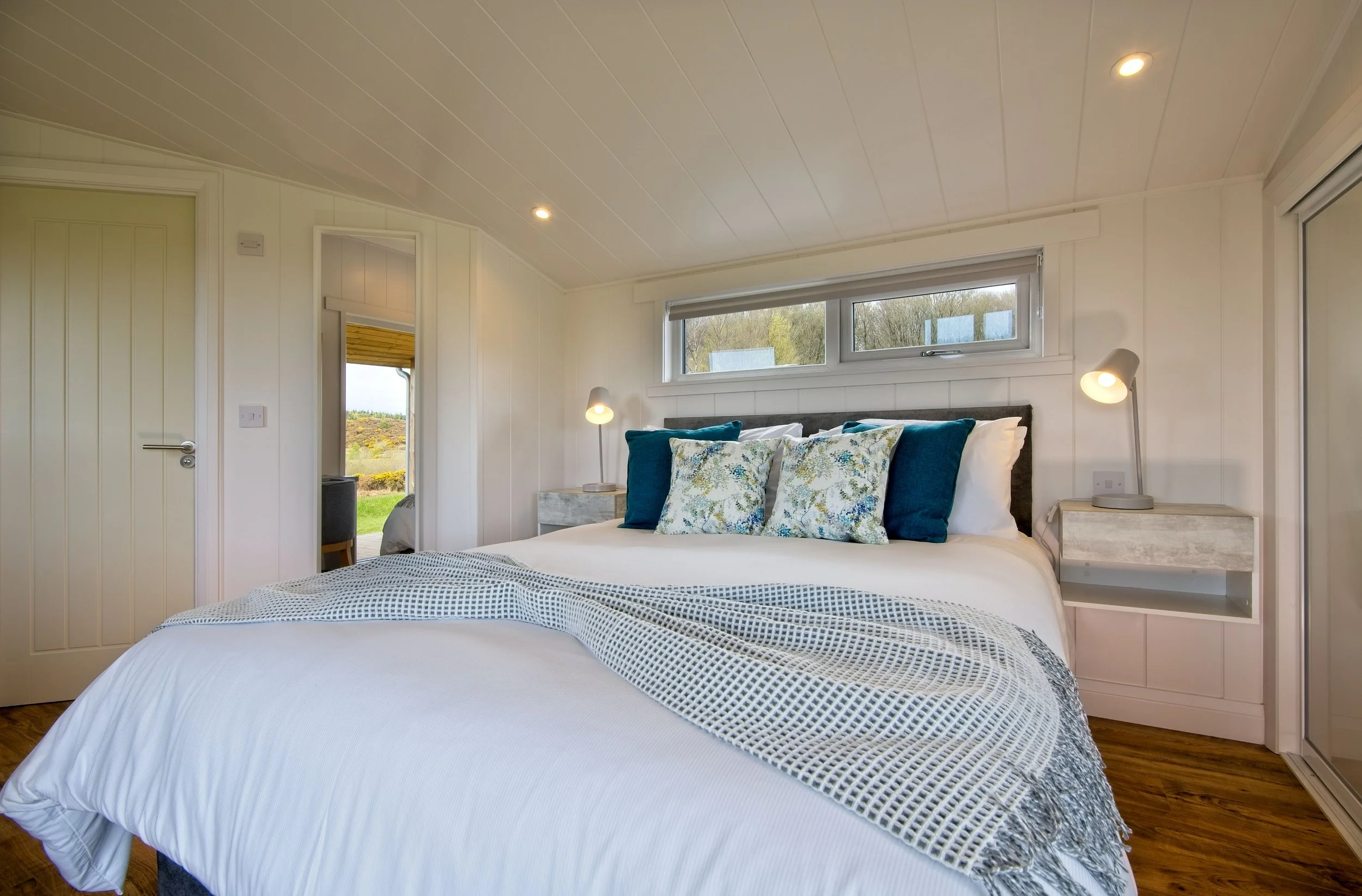 A bedroom with a neatly made white bed, blue and floral pillows, wall-mounted lamps, and a large window overlooking greenery.