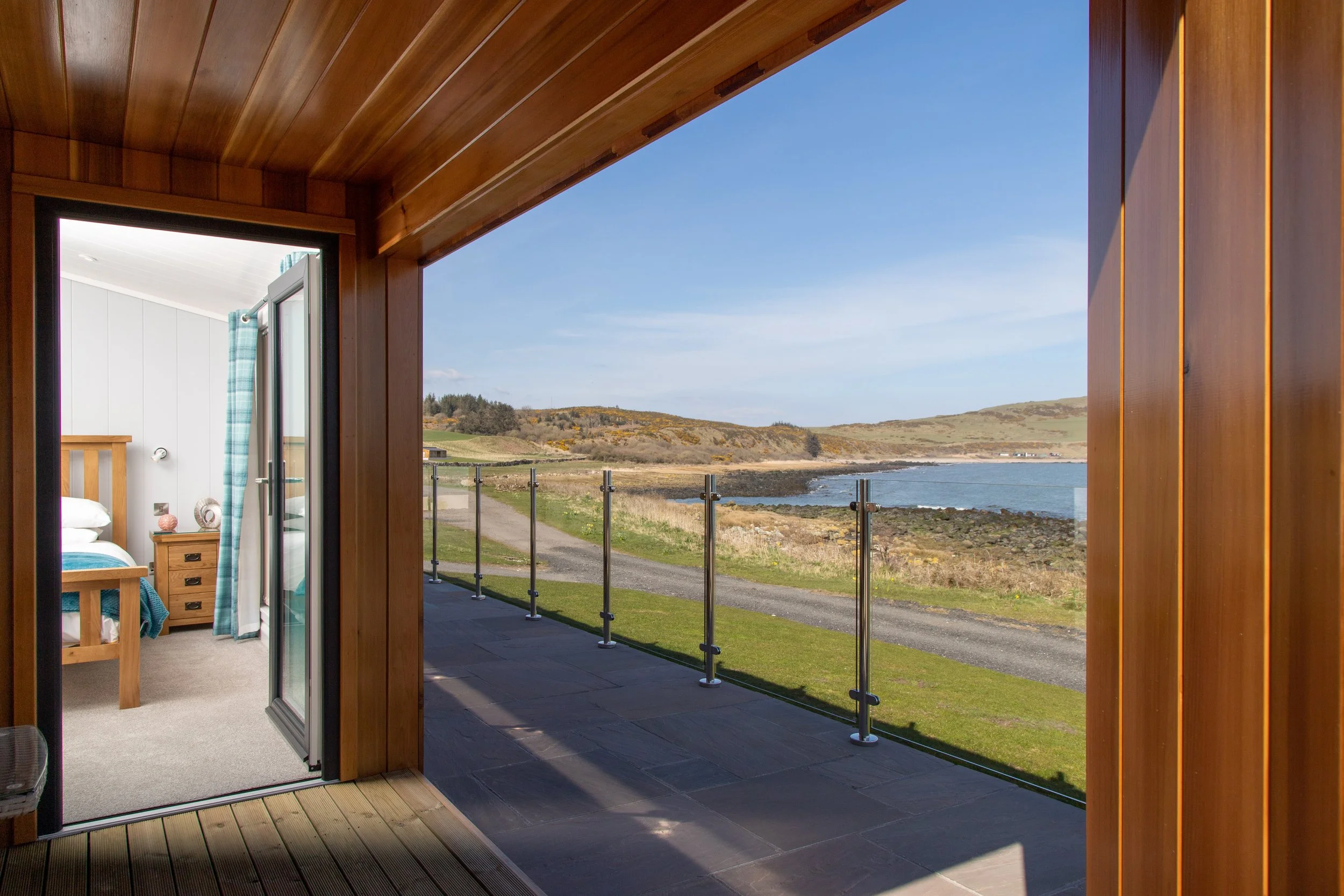 View from a balcony showing a coastal landscape with rolling hills, a rocky shoreline, and calm sea water under a clear blue sky; part of a bedroom with wooden walls and a sliding glass door.