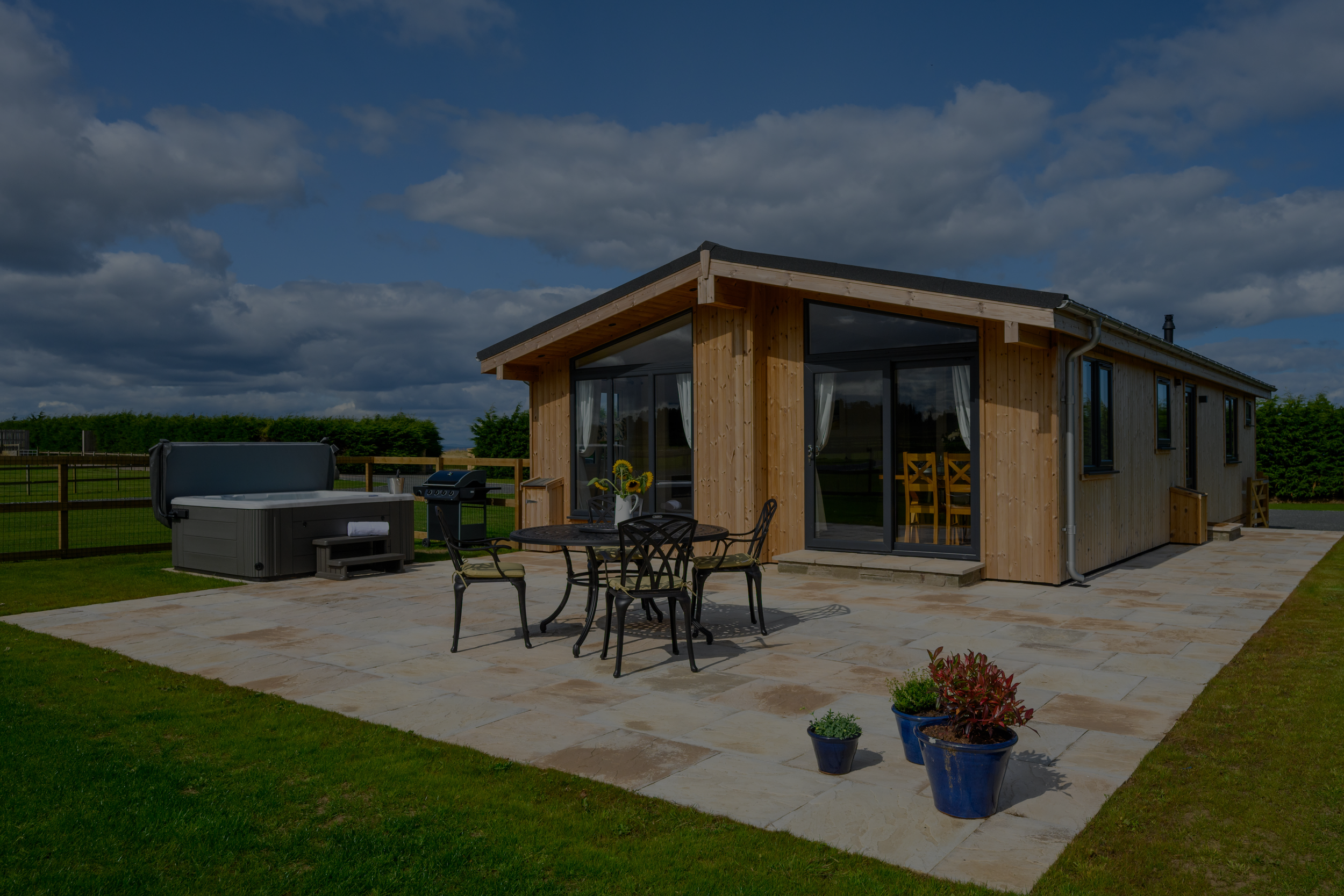Modern wooden house with large sliding glass doors, outdoor dining table with chairs, potted plants, hot tub, and a patio on a cloudy day in a rural setting.