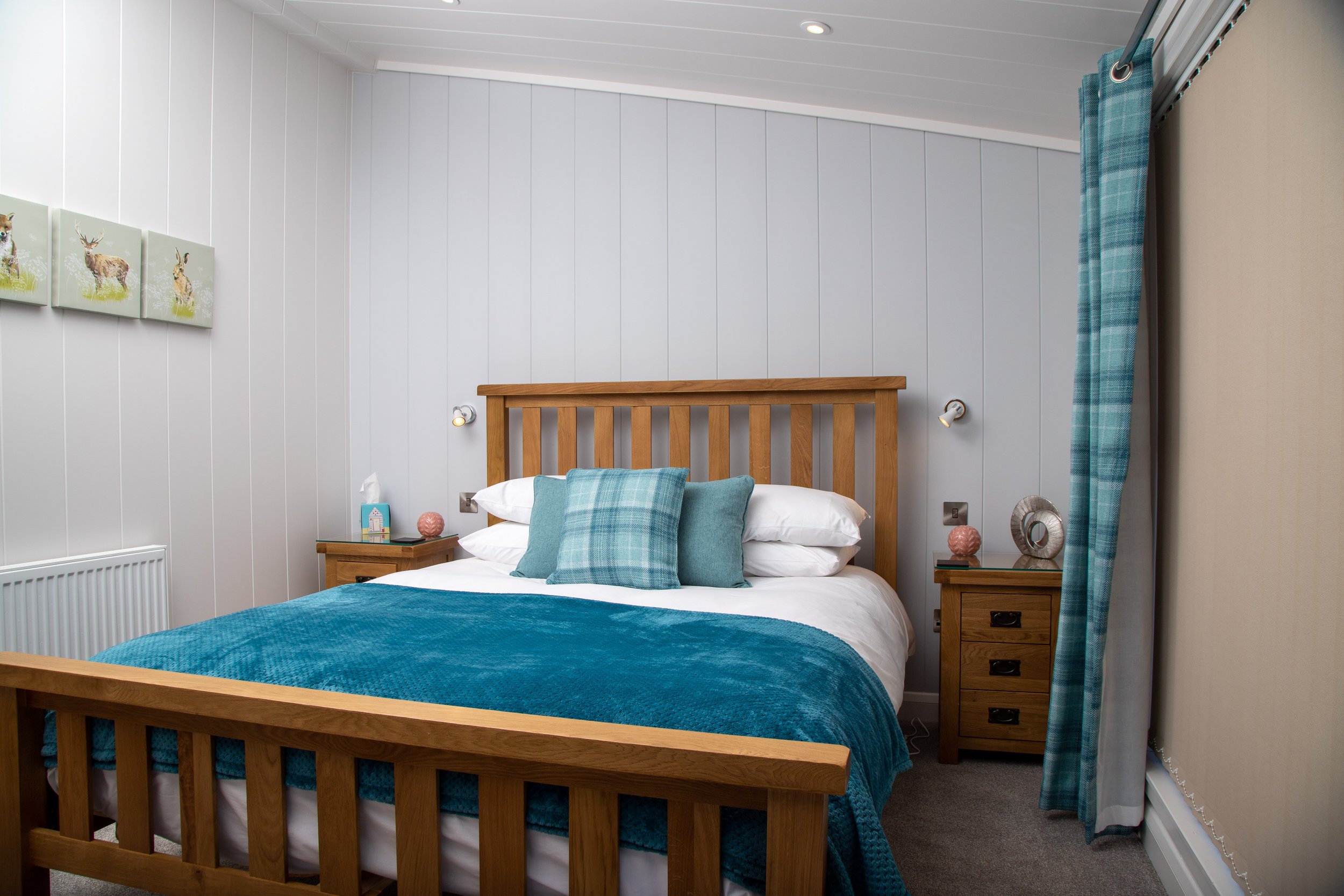 A bedroom with a wooden bed frame, white bedding, teal pillows, and a teal throw blanket. Two nightstands with lamps, decorative items, and tissues. Light-colored furniture and wall paneling, with a curtain on the right side and a wall art featuring 