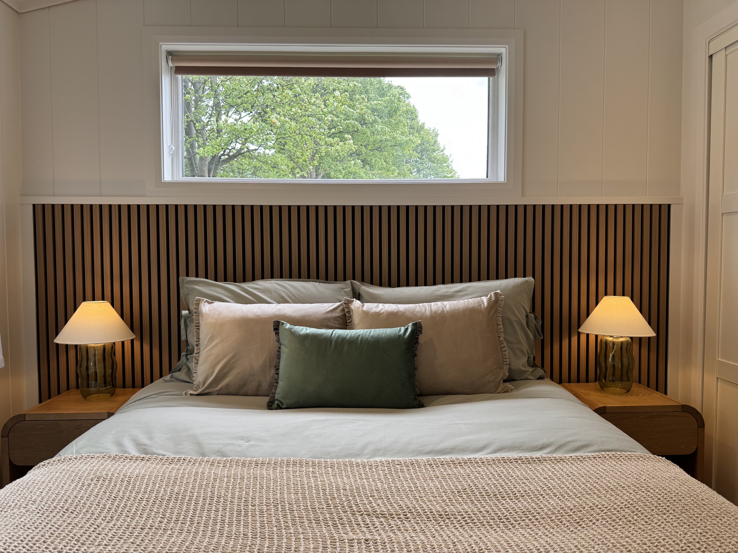 A bedroom with a bed, six pillows, two lamps on side tables, and a window showing green trees outside.
