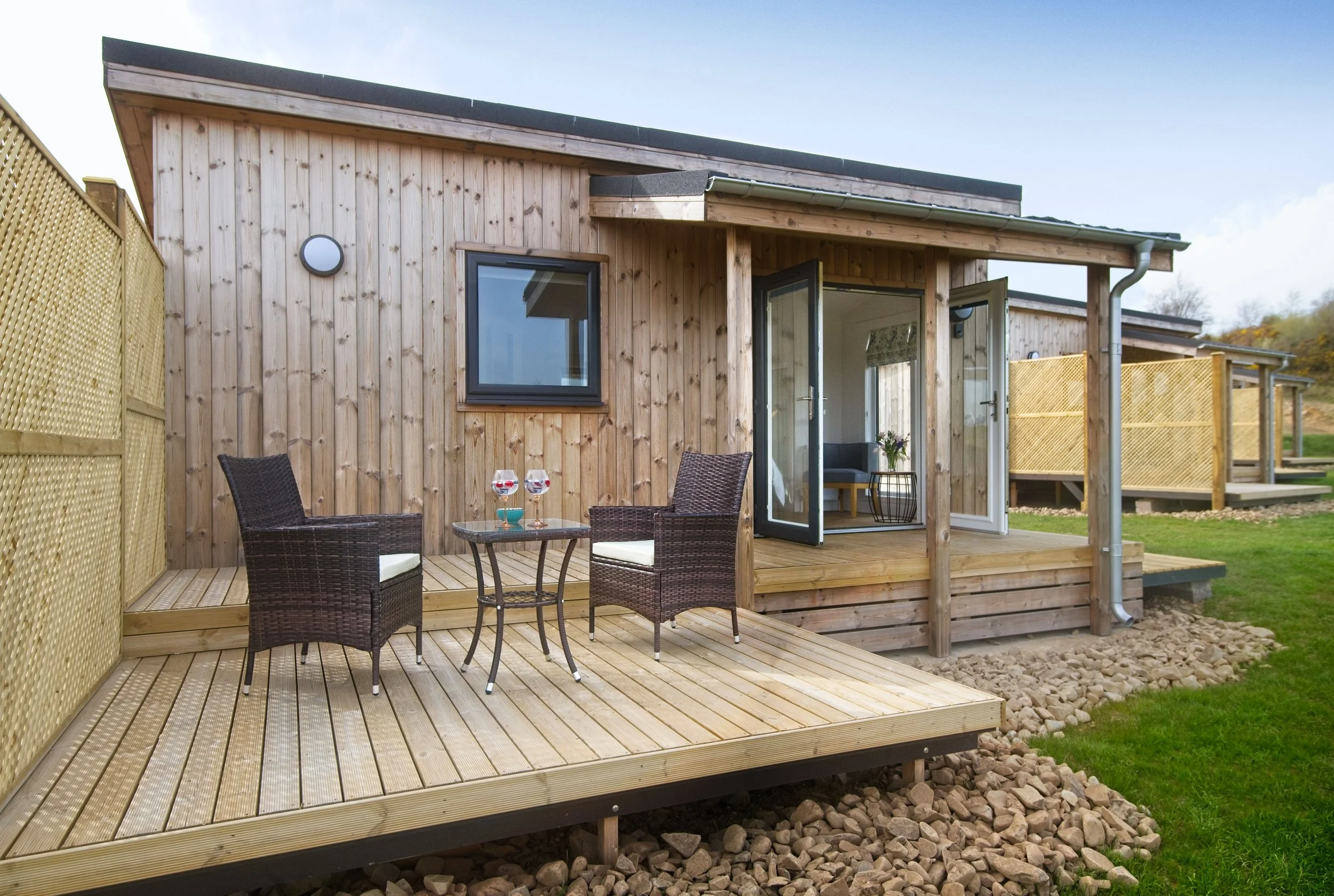 Wooden house with glass doors open to outdoor patio, two wicker chairs, small table with two glasses, grassy yard, and a neighboring wooden deck in the background.
