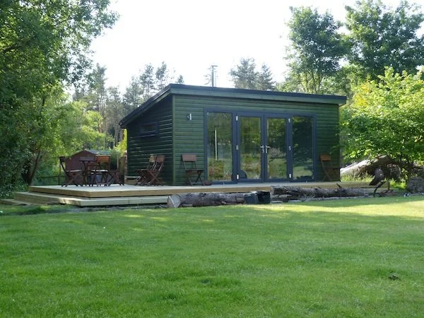 Small green cabin with large glass doors, outdoor wooden deck with chairs and table, surrounded by green grass and trees.