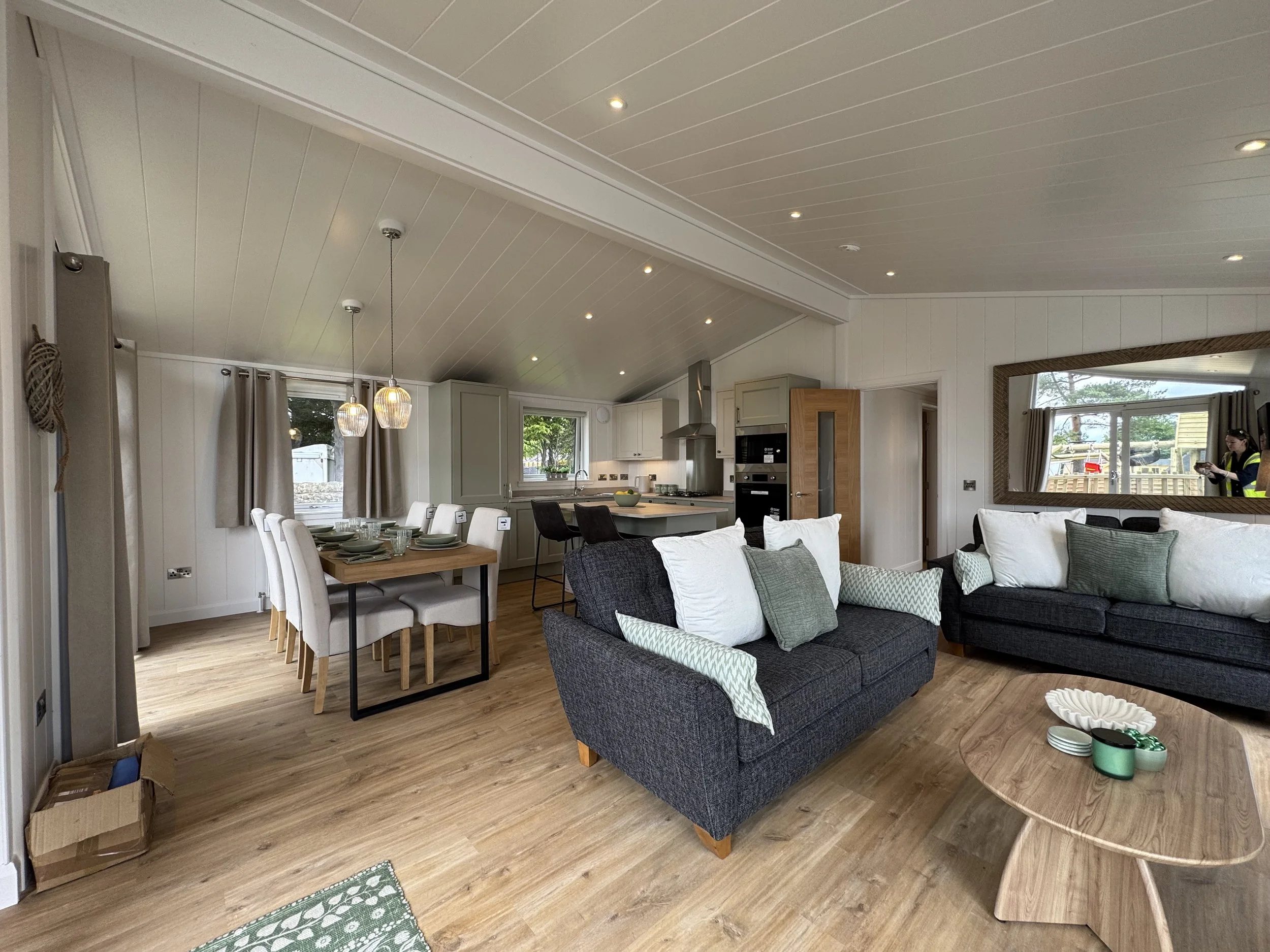 Open-concept living and dining room with a gray couch, wooden coffee table, dining table with chairs, and a kitchen in the background, featuring white cabinetry and a breakfast bar.
