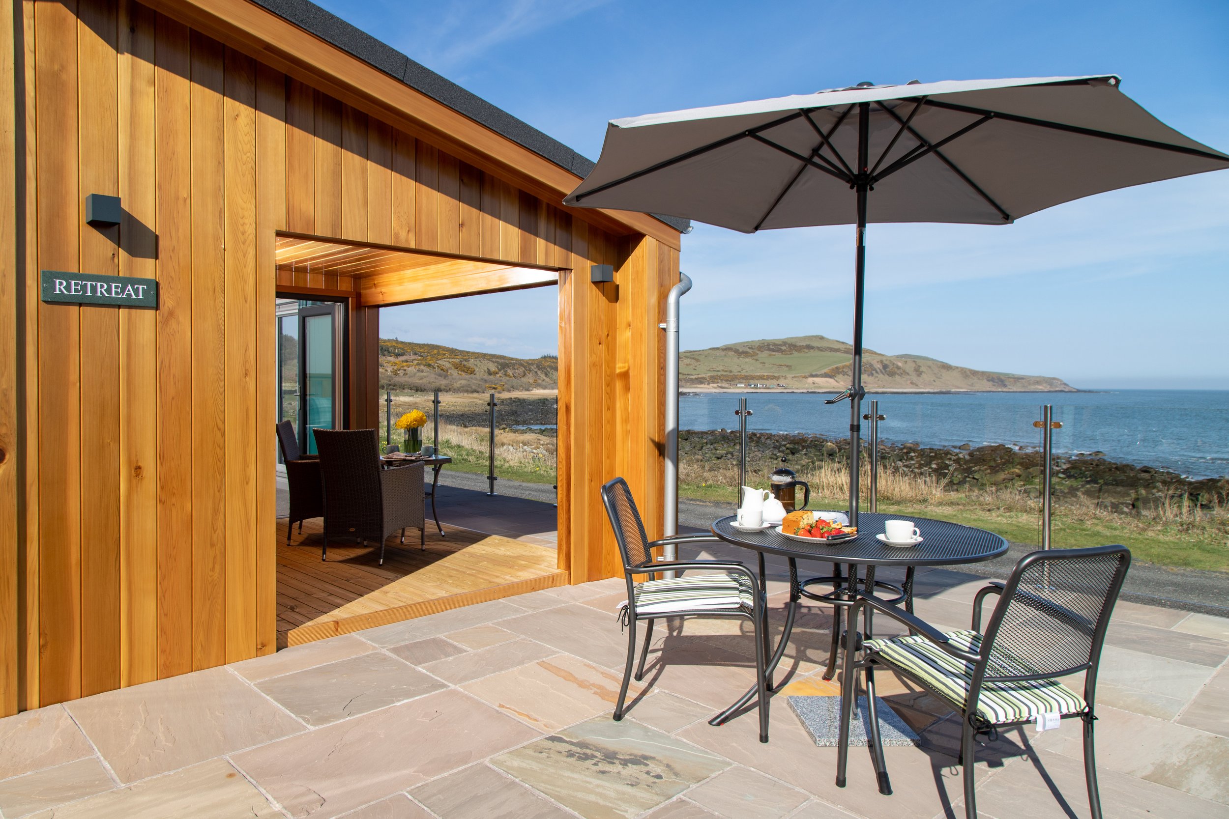 Outdoor patio with a round table and three chairs under a large umbrella, overlooking a shoreline with water and distant hills, adjacent to a wooden building labeled 'Retreat'.