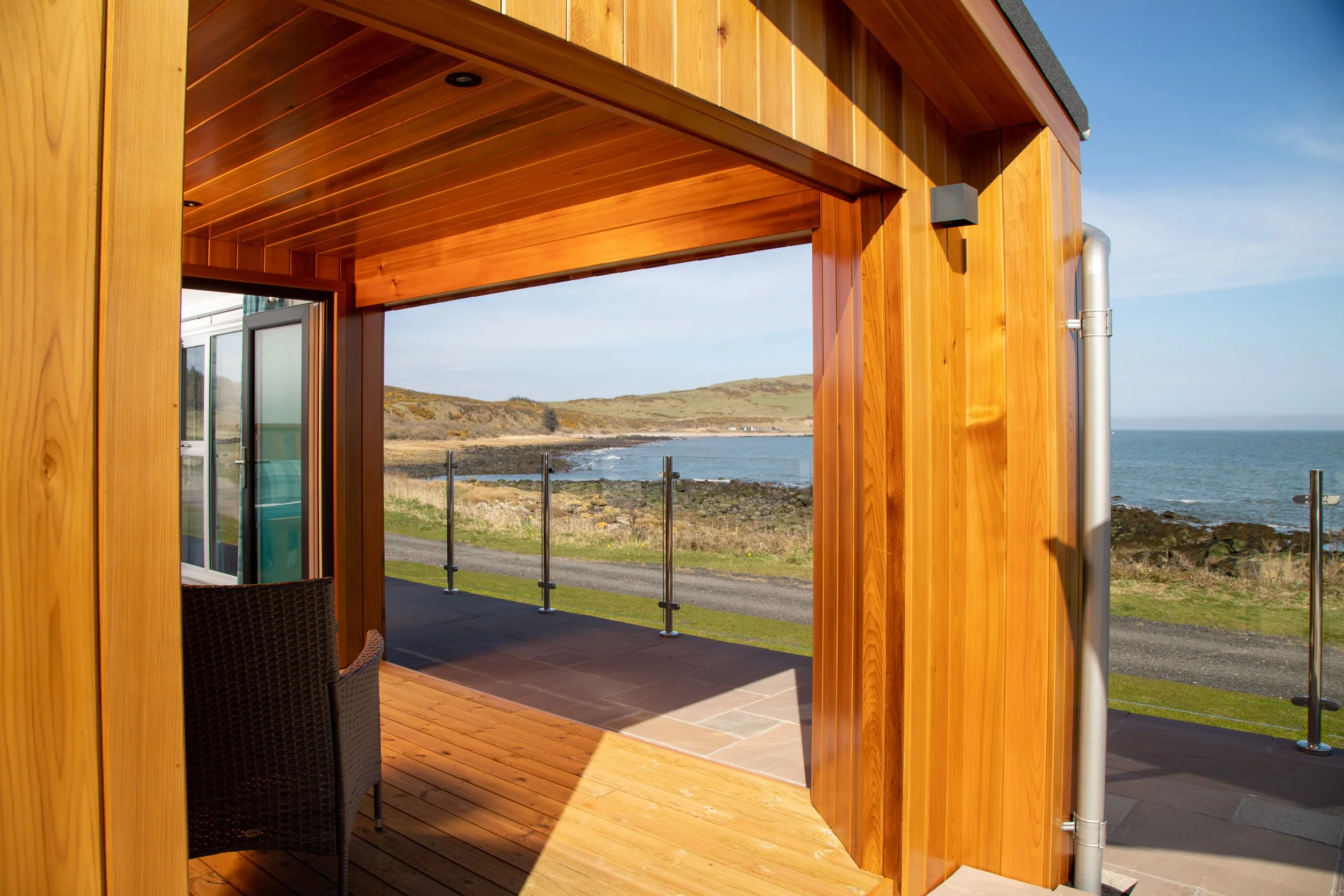 View from a wooden balcony overlooking a coastline with grassy hills, rocky shoreline, and the ocean on a clear day.