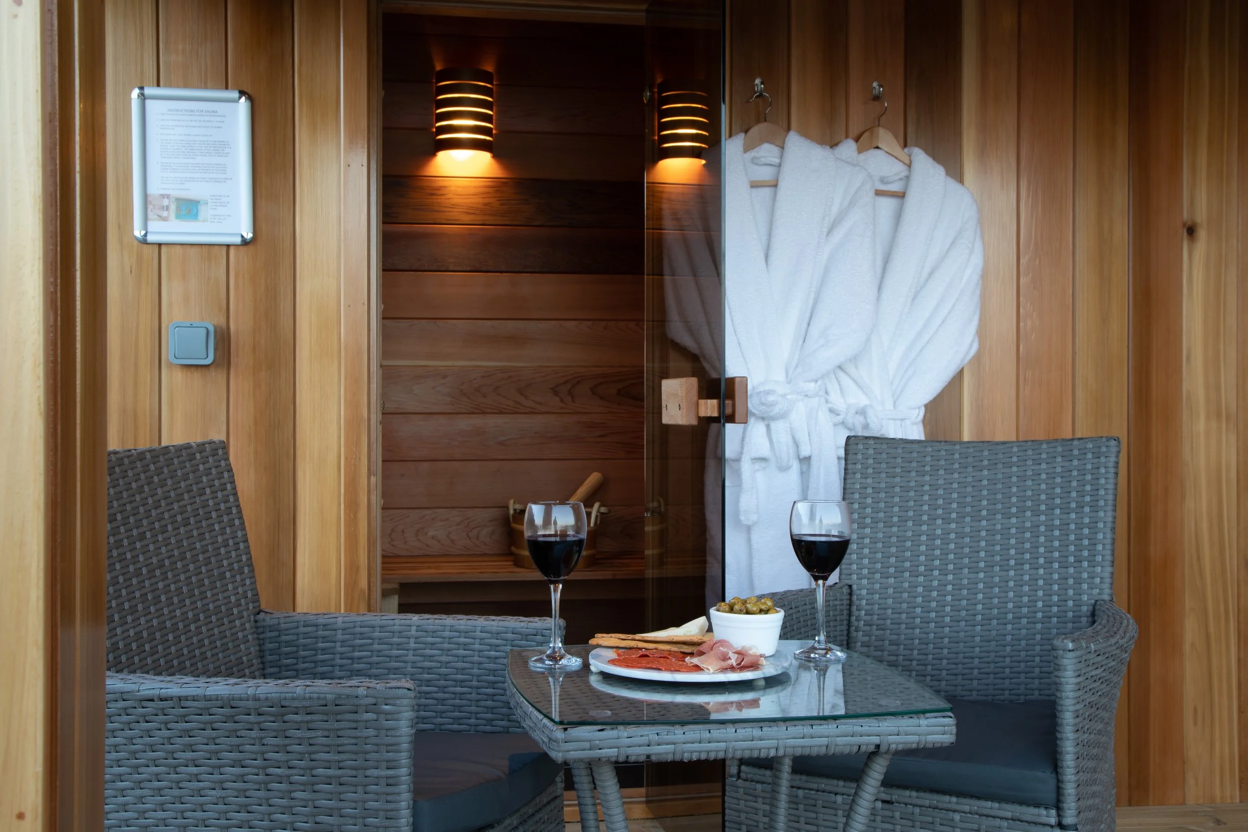 A cozy indoor setting with a small glass-top table, two wicker chairs, and a plate of charcuterie with two glasses of red wine. In the background, wooden walls and a glass door reveal a sauna with white bathrobes hanging on hooks.